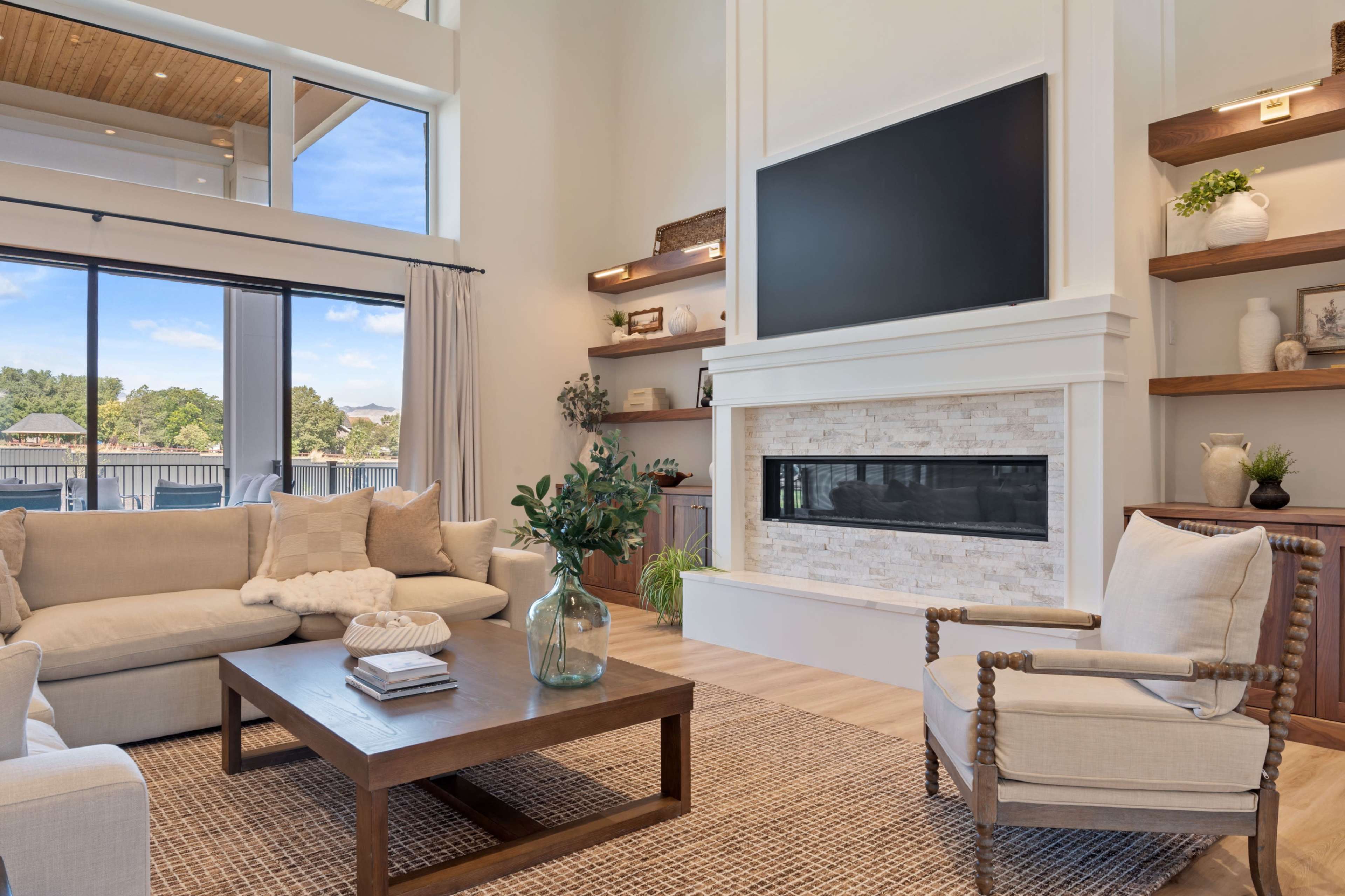A modern living room features a large television above a stone fireplace, surrounded by light-colored furniture and large windows that provide ample natural light.
