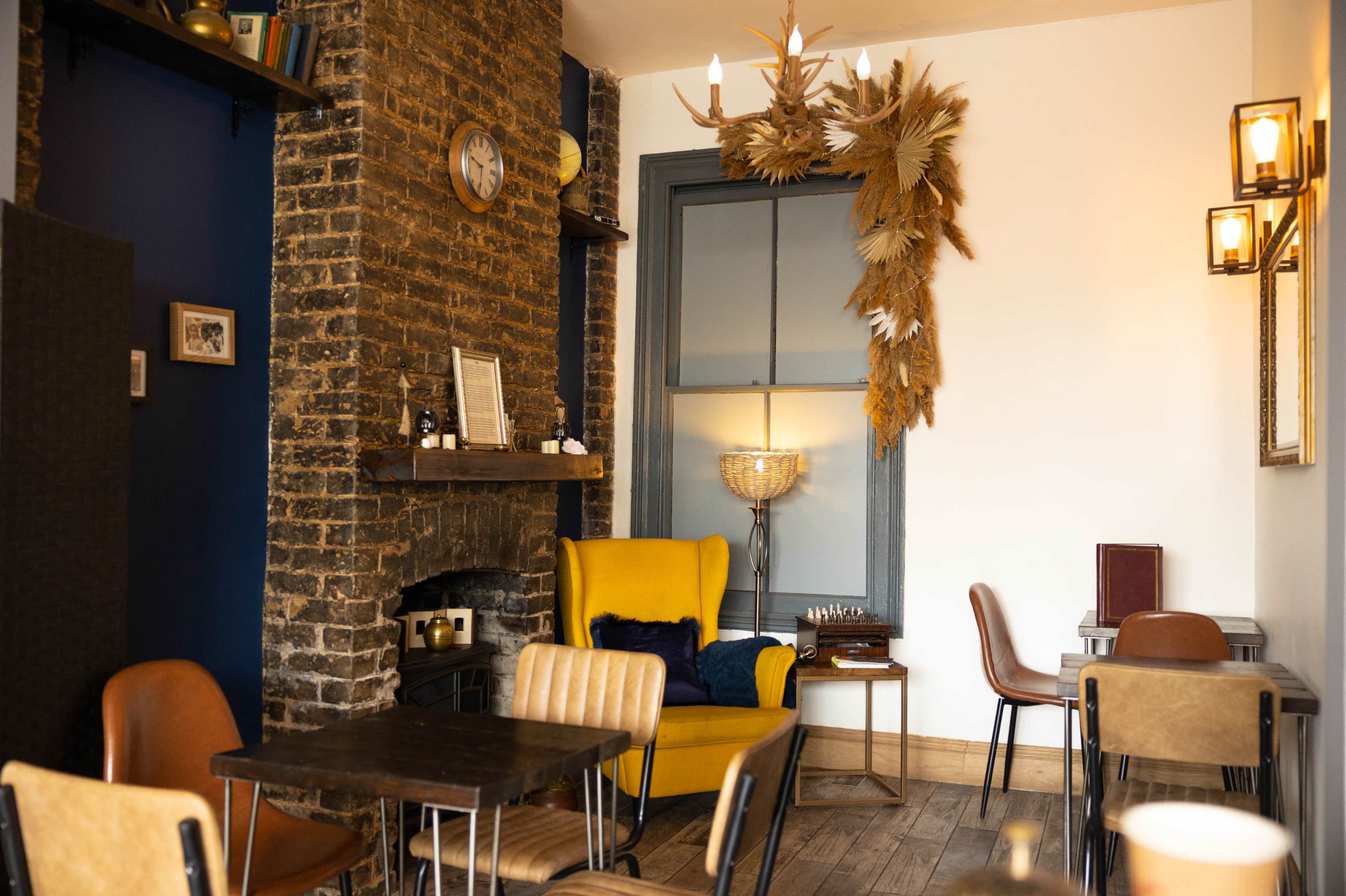 A cozy café interior with a yellow armchair, wooden tables, and a decorative wall featuring a large arrangement of dried plants above a fireplace.