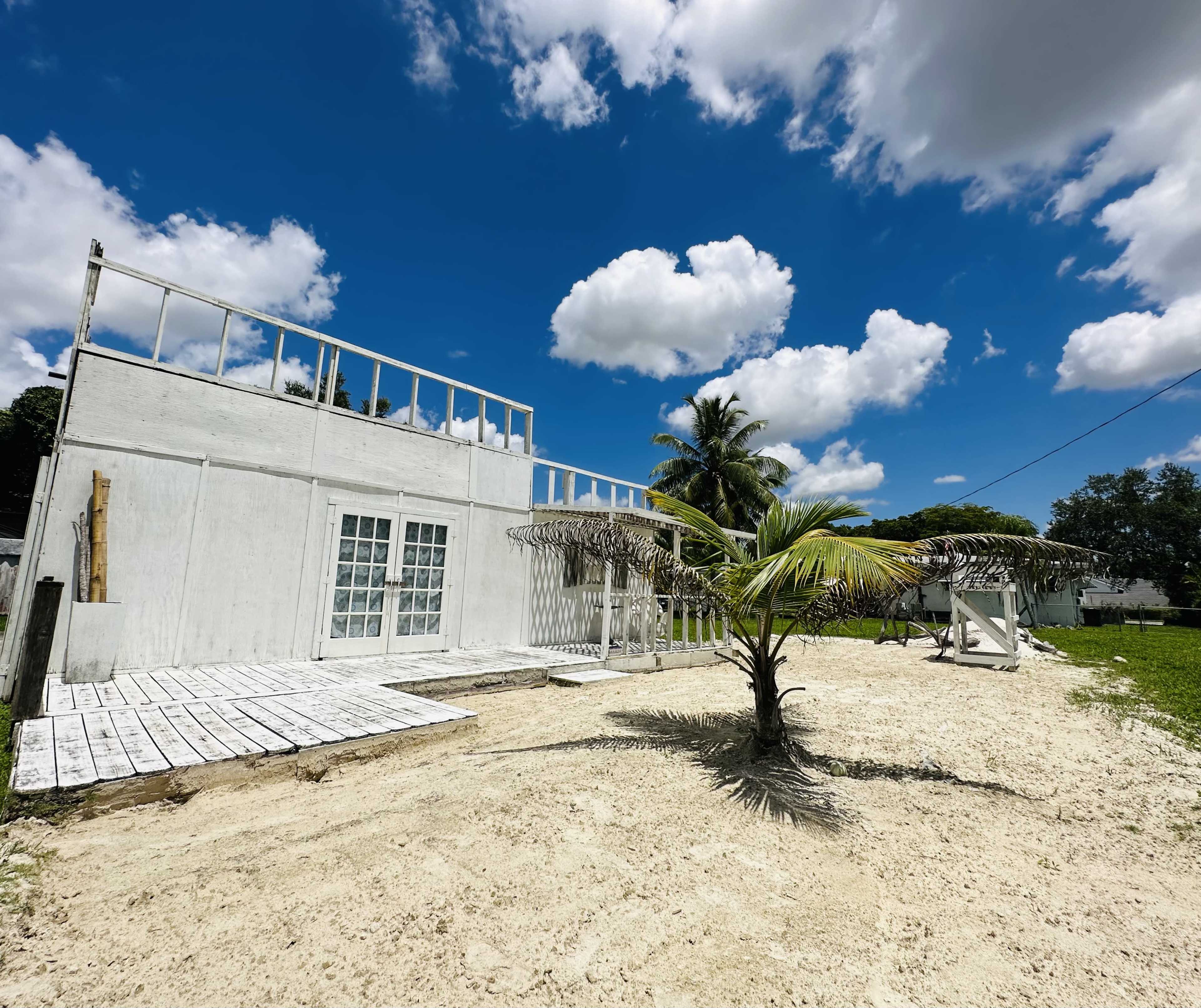 A white building with a wooden deck and an umbrella structure stands next to a palm tree on a sandy area under a bright blue sky with clouds.