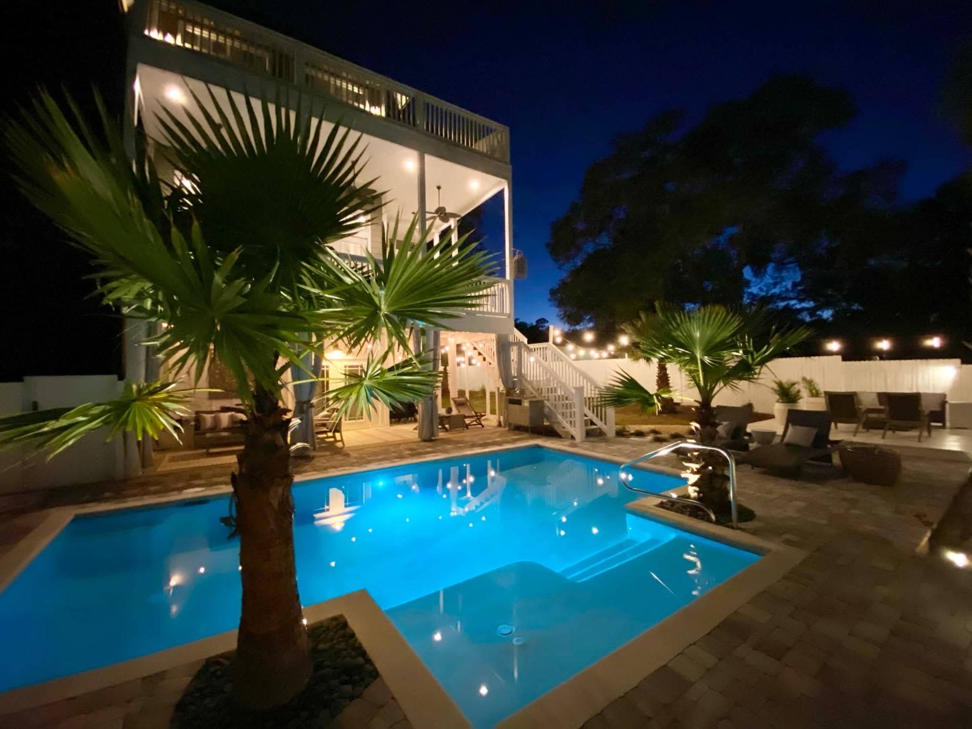 A two-story house with a patio and pool is illuminated at night, surrounded by palm trees and decorative string lights.