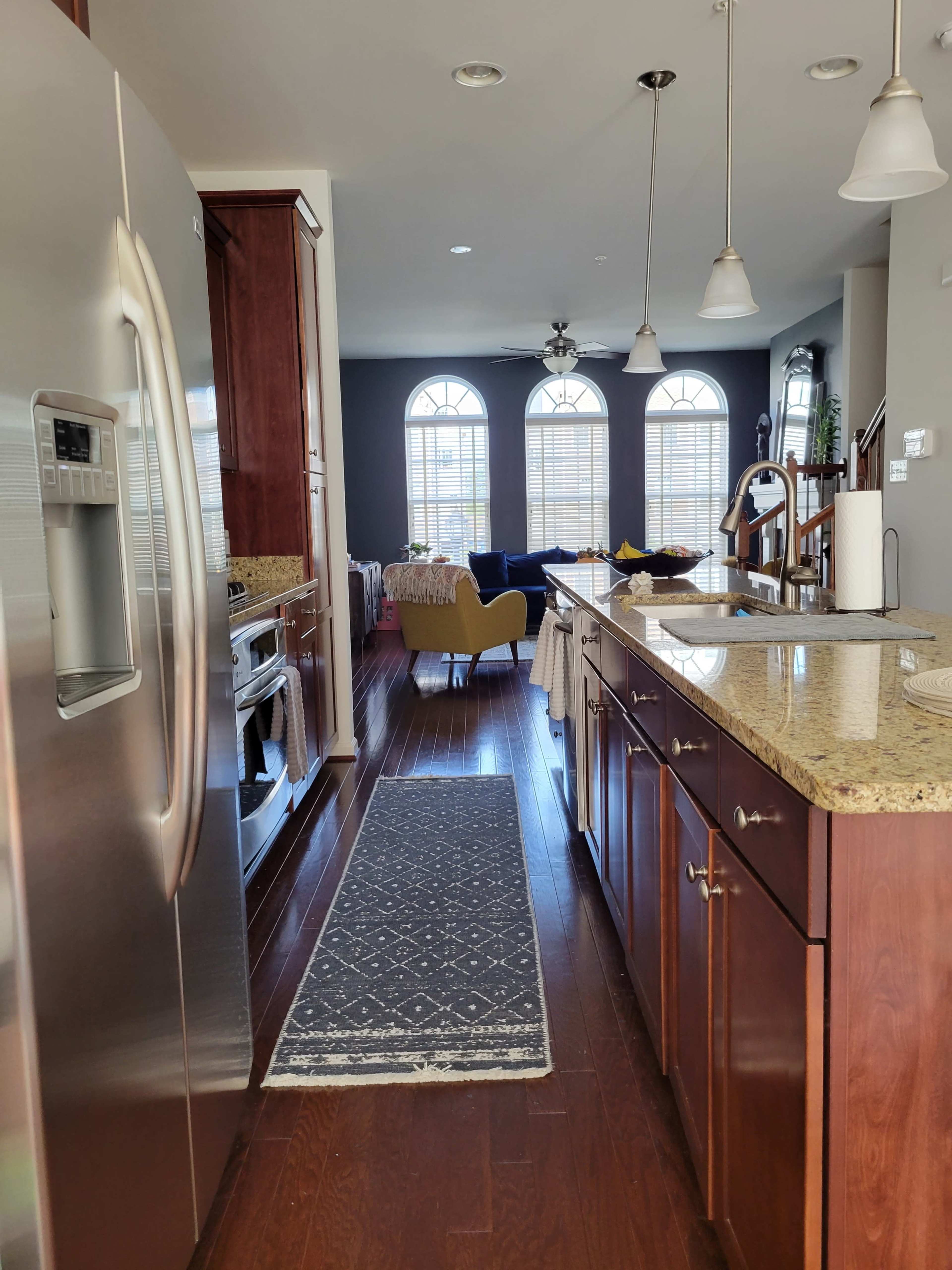 A kitchen with dark wood cabinetry, a stainless steel refrigerator, and a patterned rug running along the floor, leading to a living area with large windows and a sofa in the background.