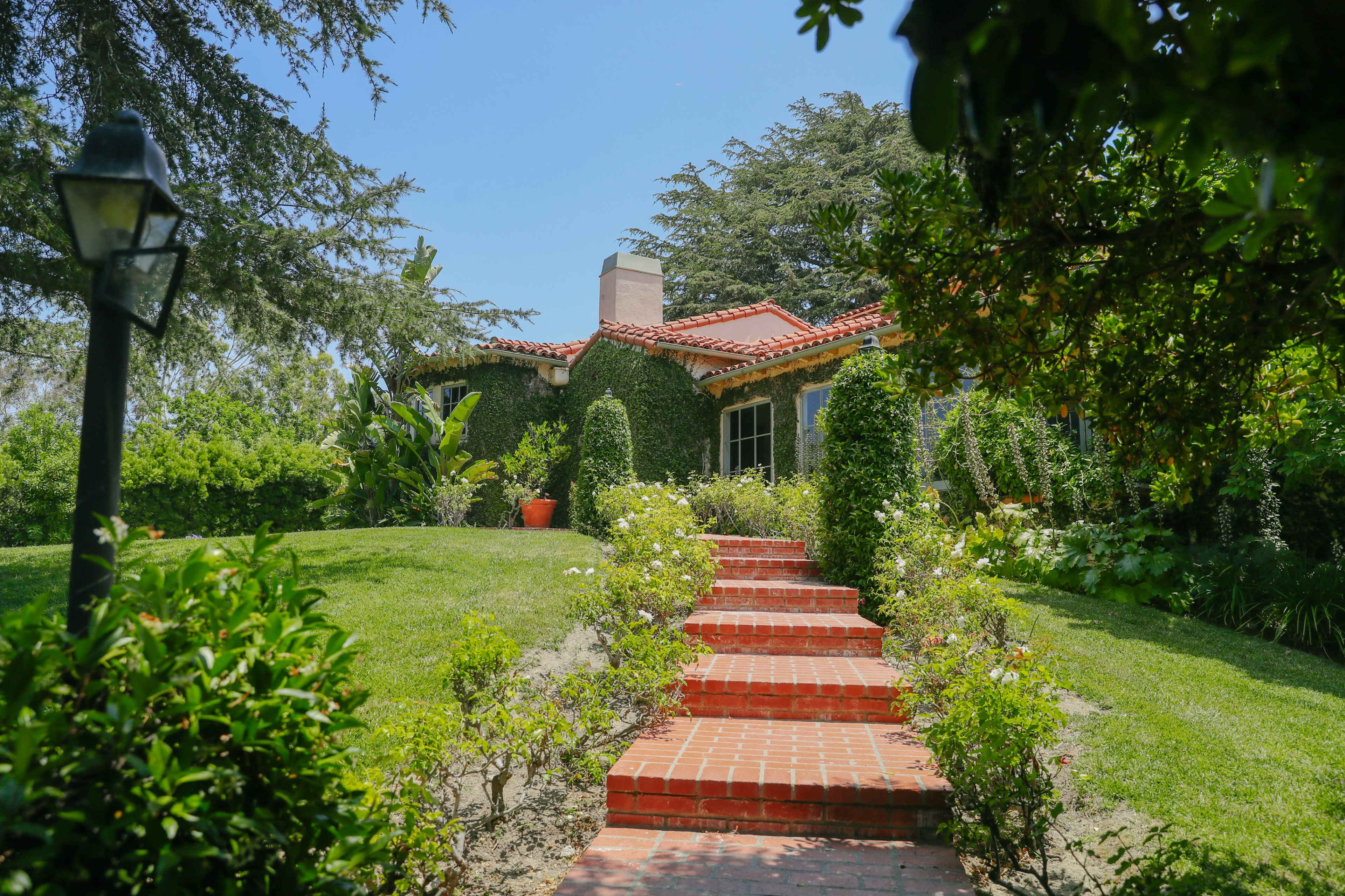 A brick pathway leads to a charming house surrounded by lush greenery and hedges.