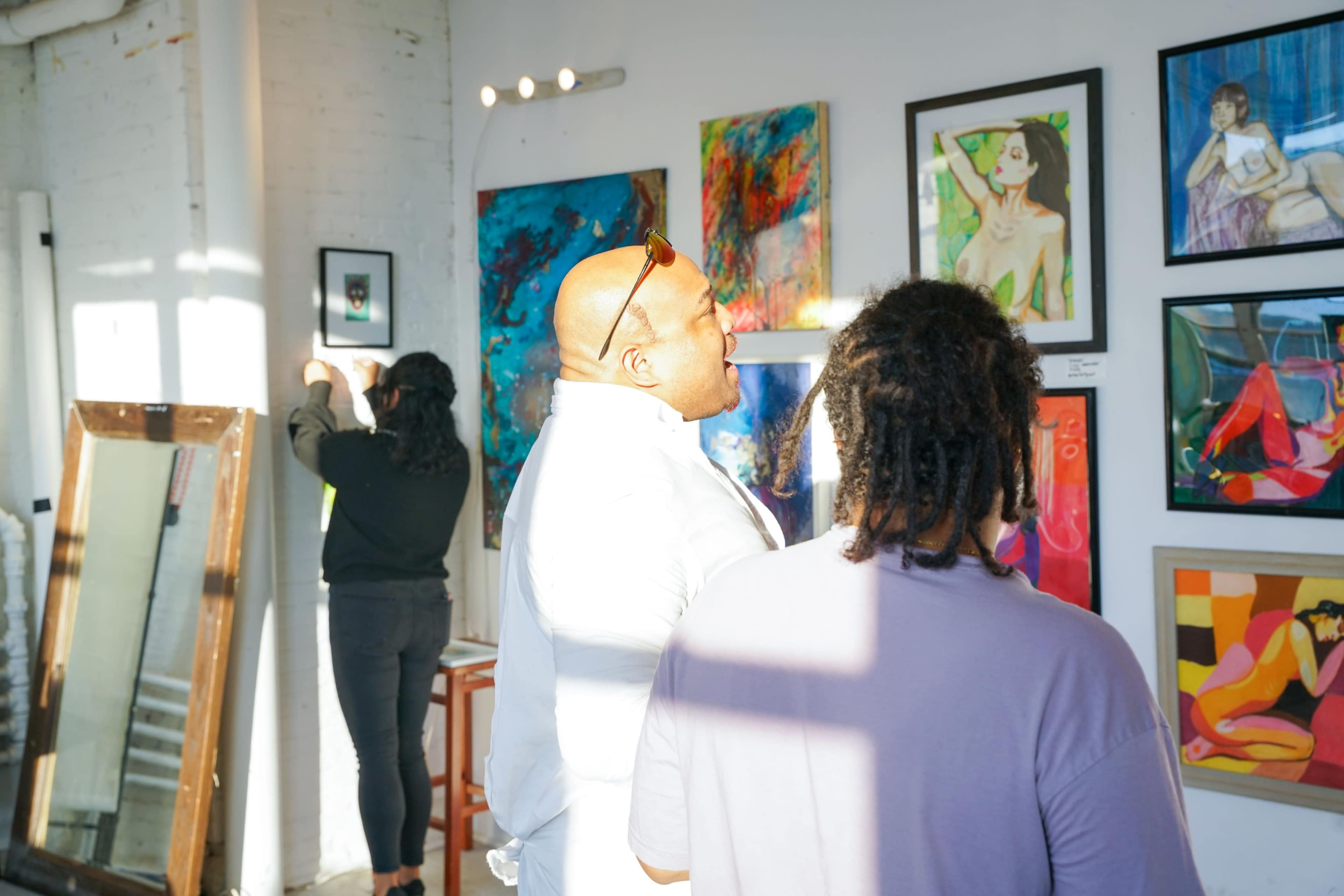 In an art gallery, a man talks while two other individuals examine colorful paintings on the walls.