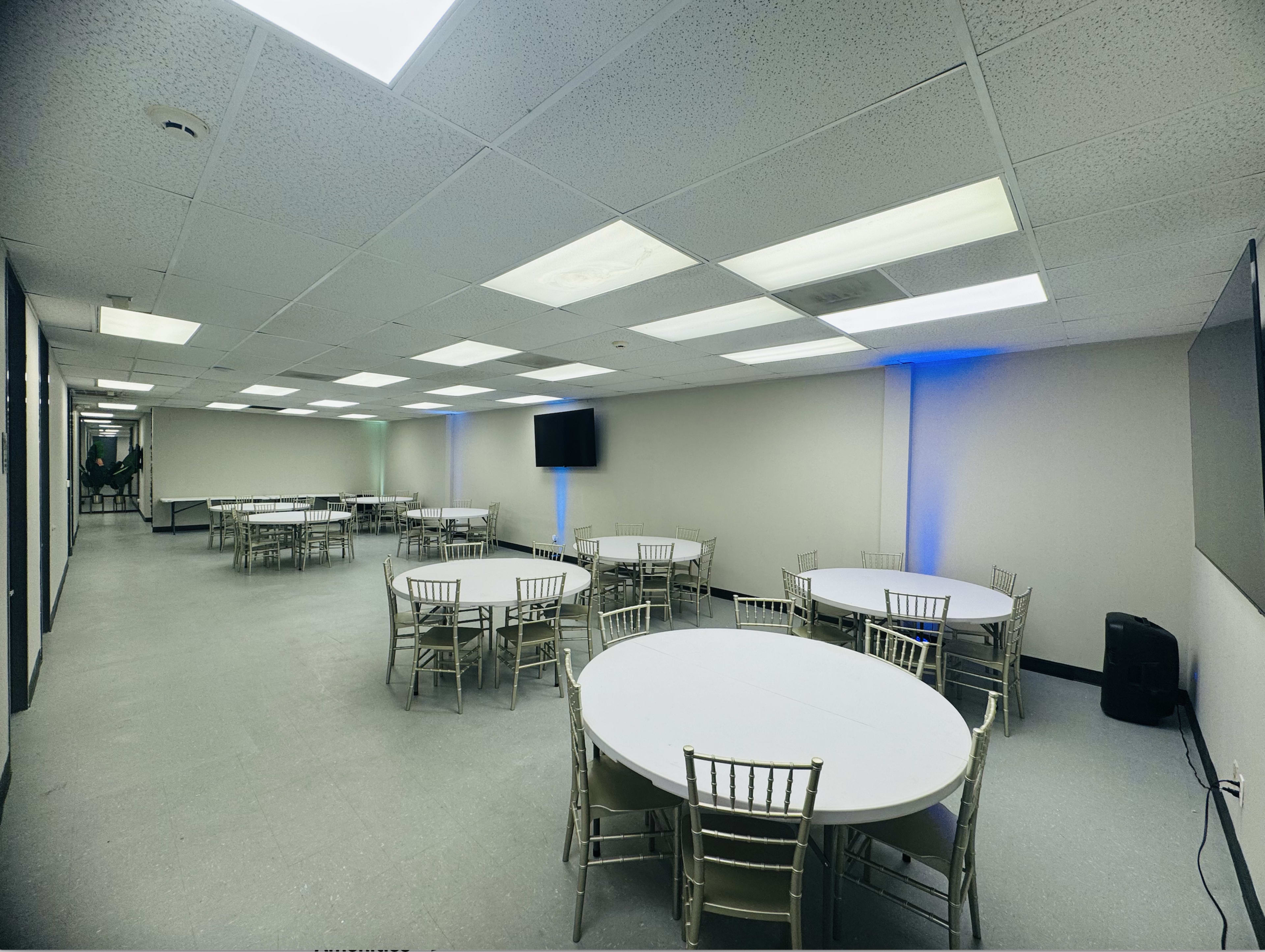 A spacious room features multiple round tables with chairs arranged around them, illuminated by overhead lights.