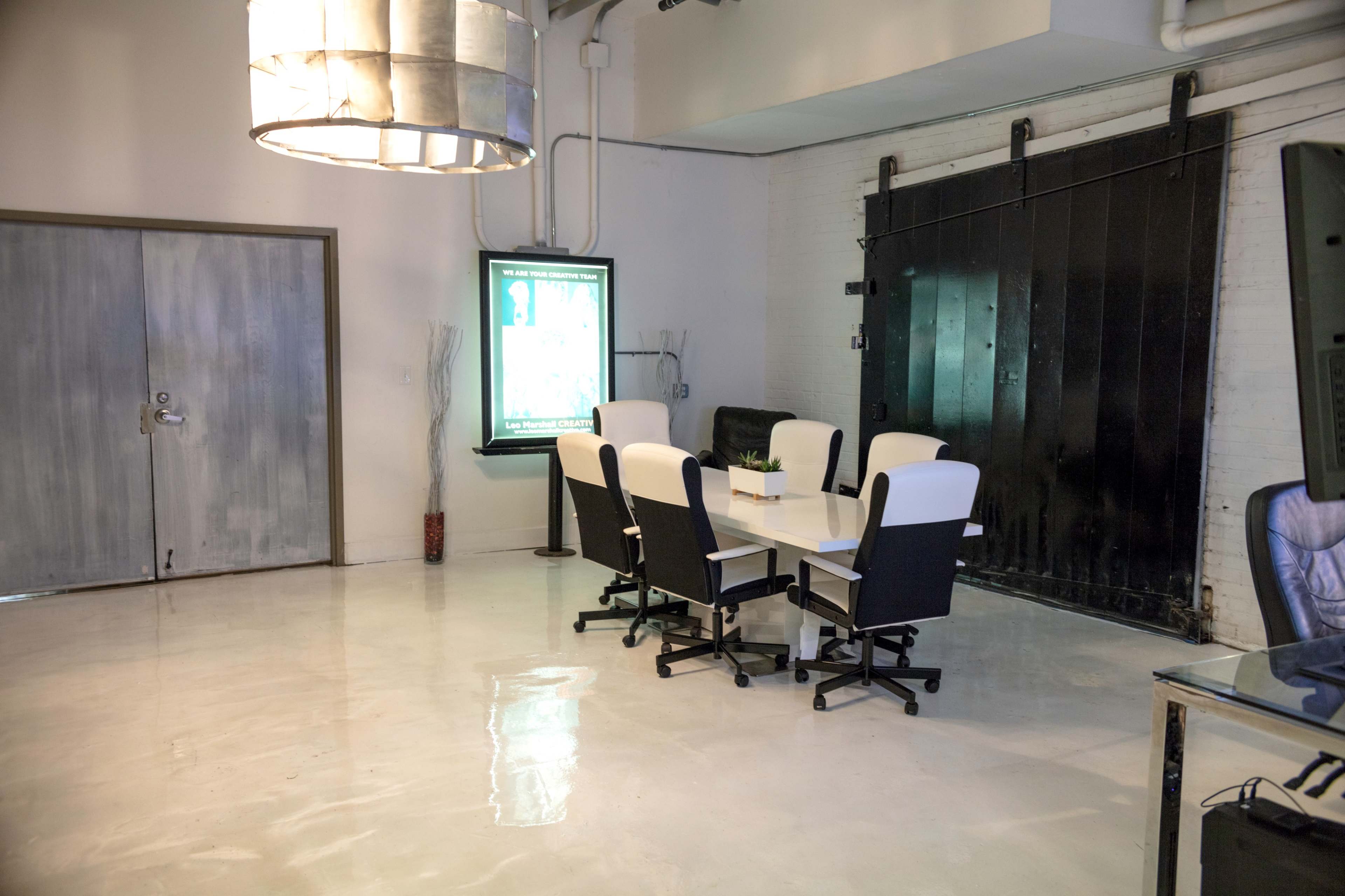 A meeting room with a long table surrounded by six chairs, a large screen displaying content, and a black door in the background.