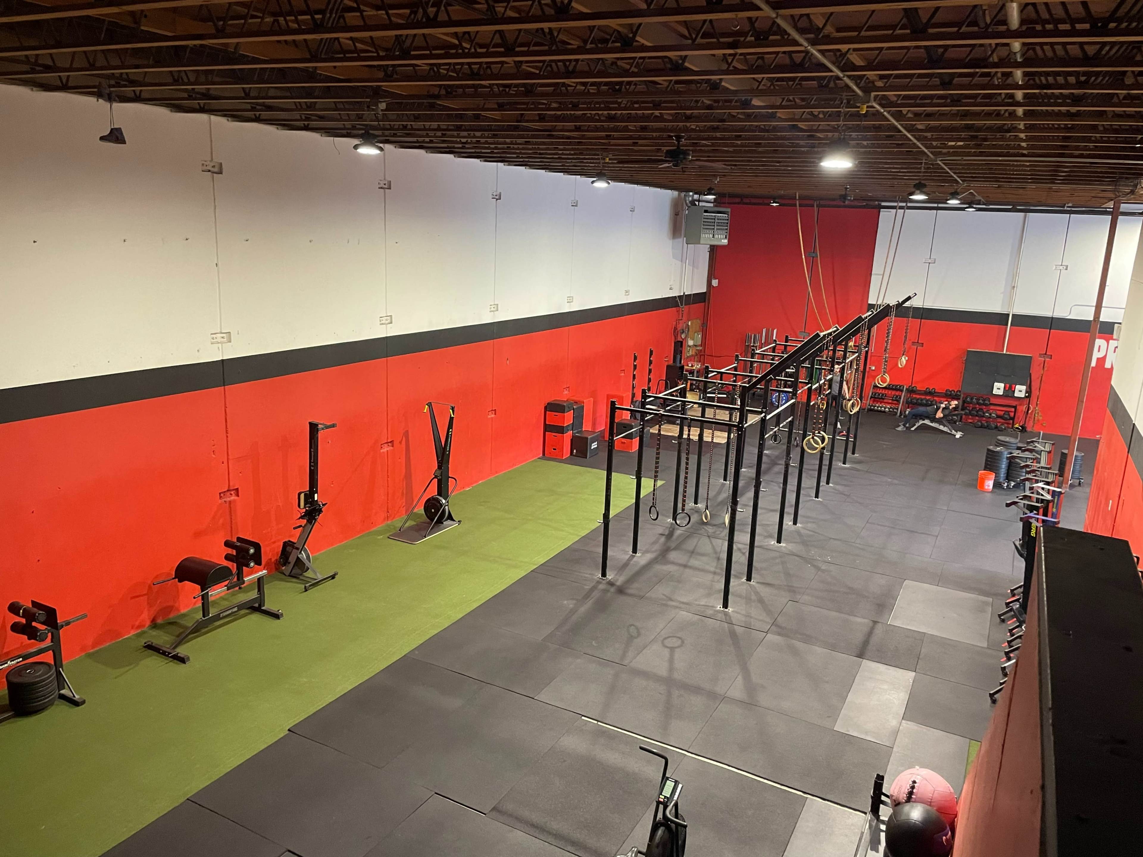 A spacious gym with red walls, black equipment, and various workout stations arranged on a green and black rubber floor.