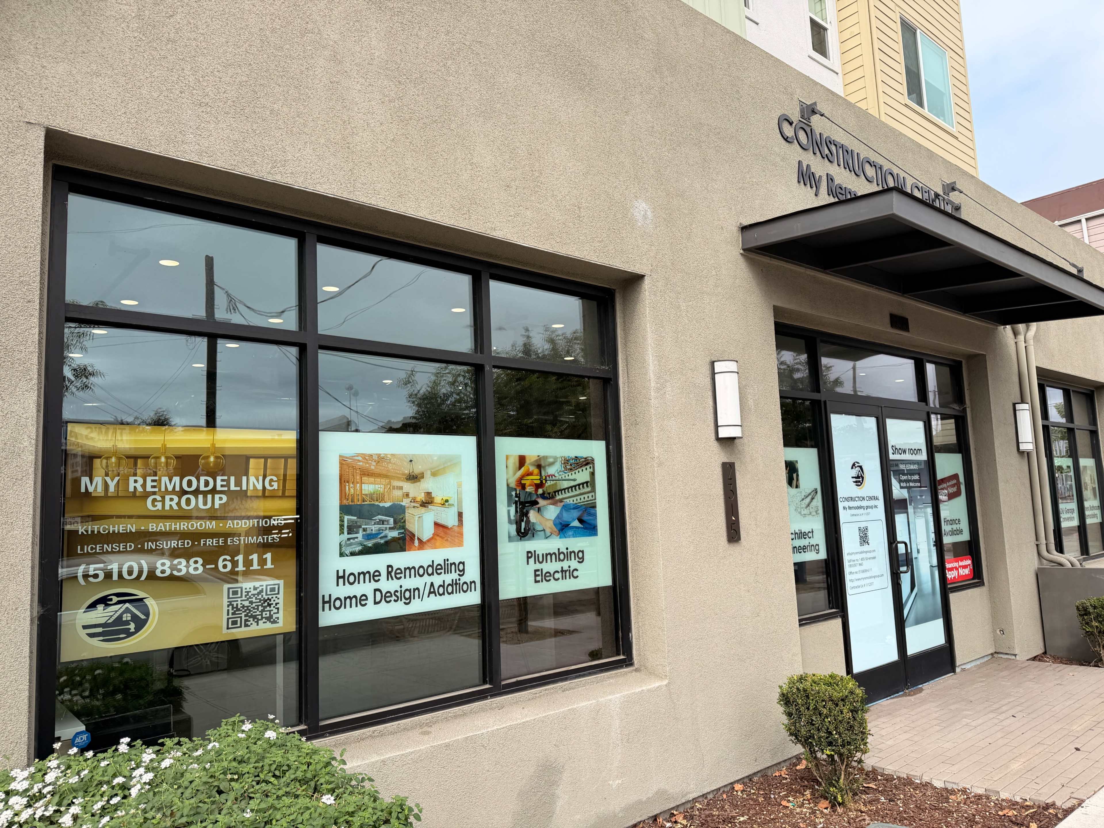 The image shows the exterior of a construction and remodeling business with large glass windows displaying advertisements for services such as home design, plumbing, and electric work.