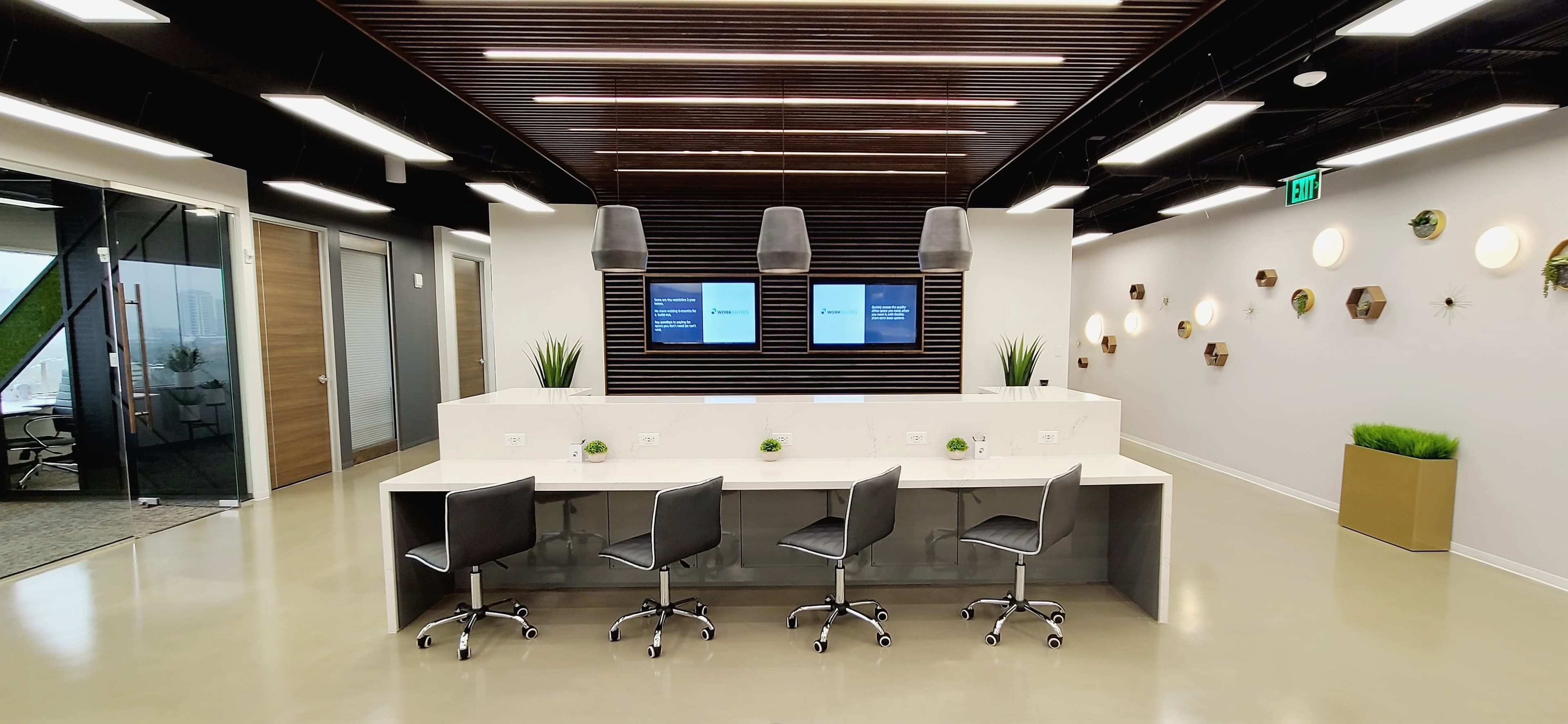 The image shows a modern office interior featuring a reception area with four black chairs at a beige counter, two screens mounted on the wall, and decorative plants on the counter and walls.