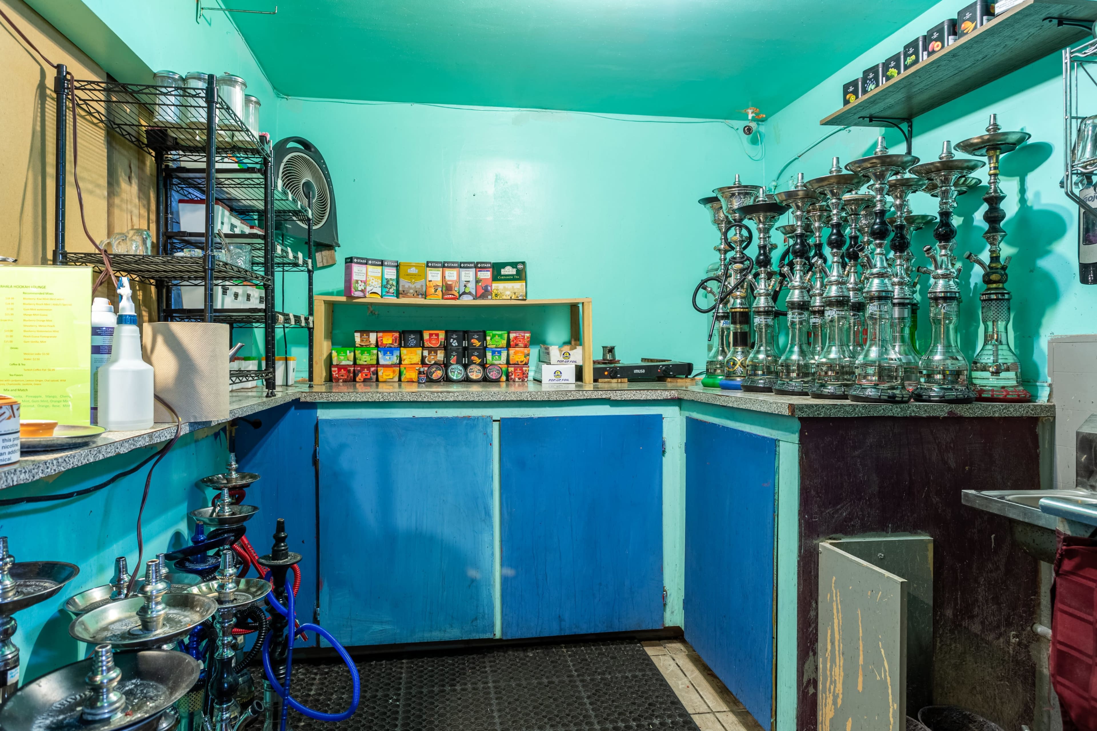 A small, turquoise-colored room with shelves containing hookahs and various flavored tobacco products arranged on a counter.