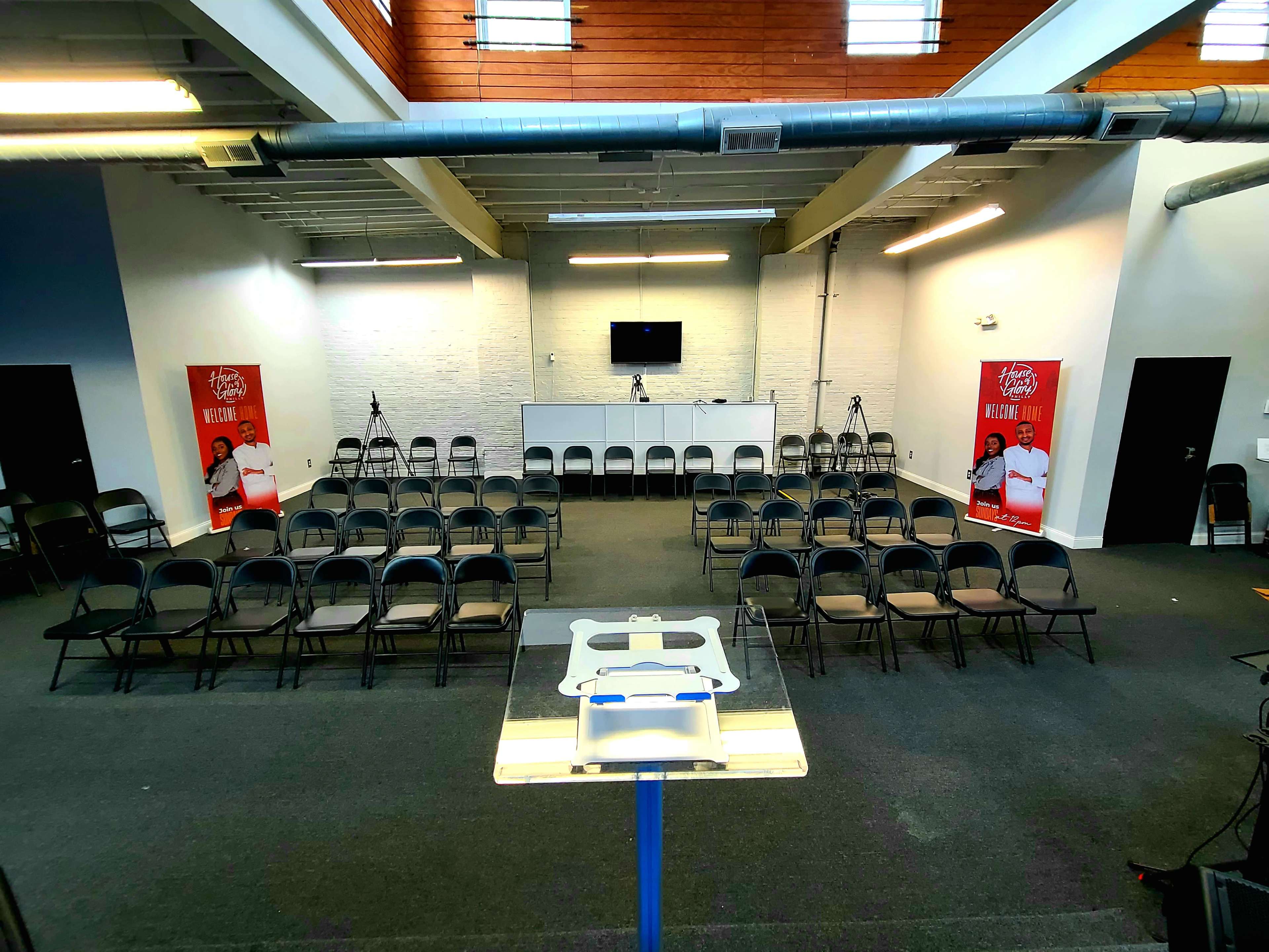 A conference room set up with rows of chairs facing a stage, flanked by two promotional banners and equipped with cameras and a monitor.