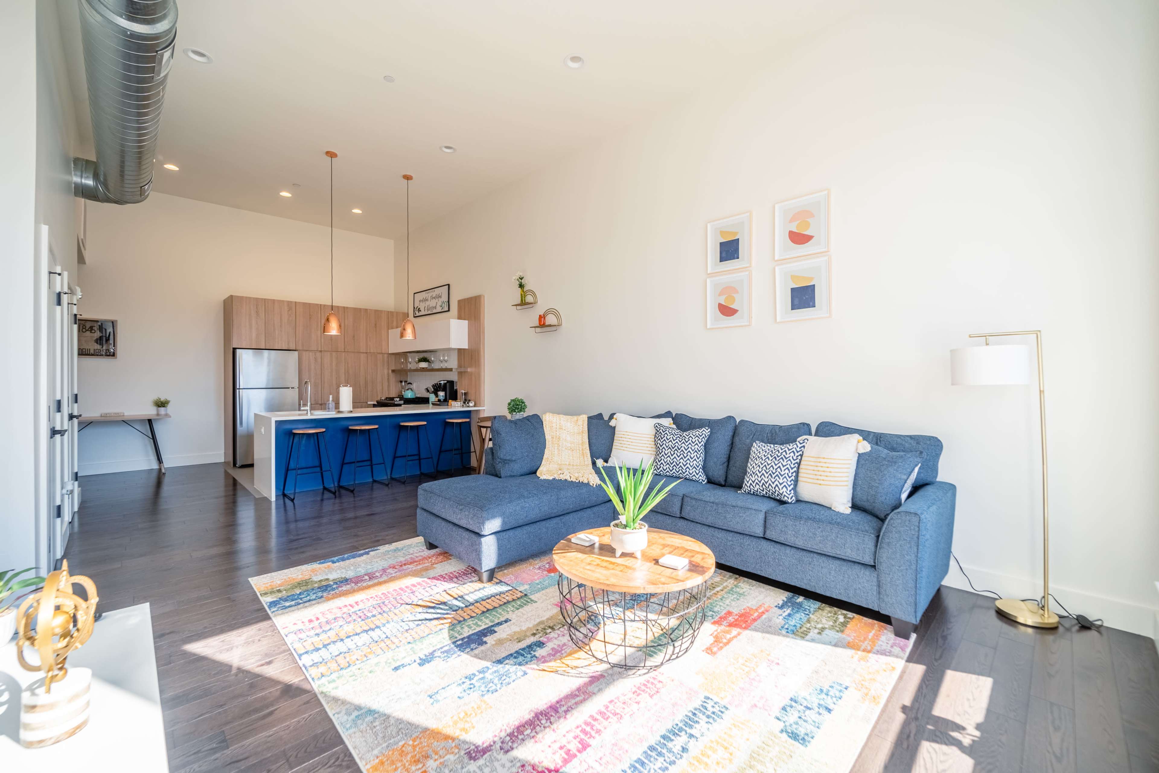 A modern living room features a blue sectional sofa, a round coffee table with plants, and a view of an open kitchen with wooden cabinets and bar seating.