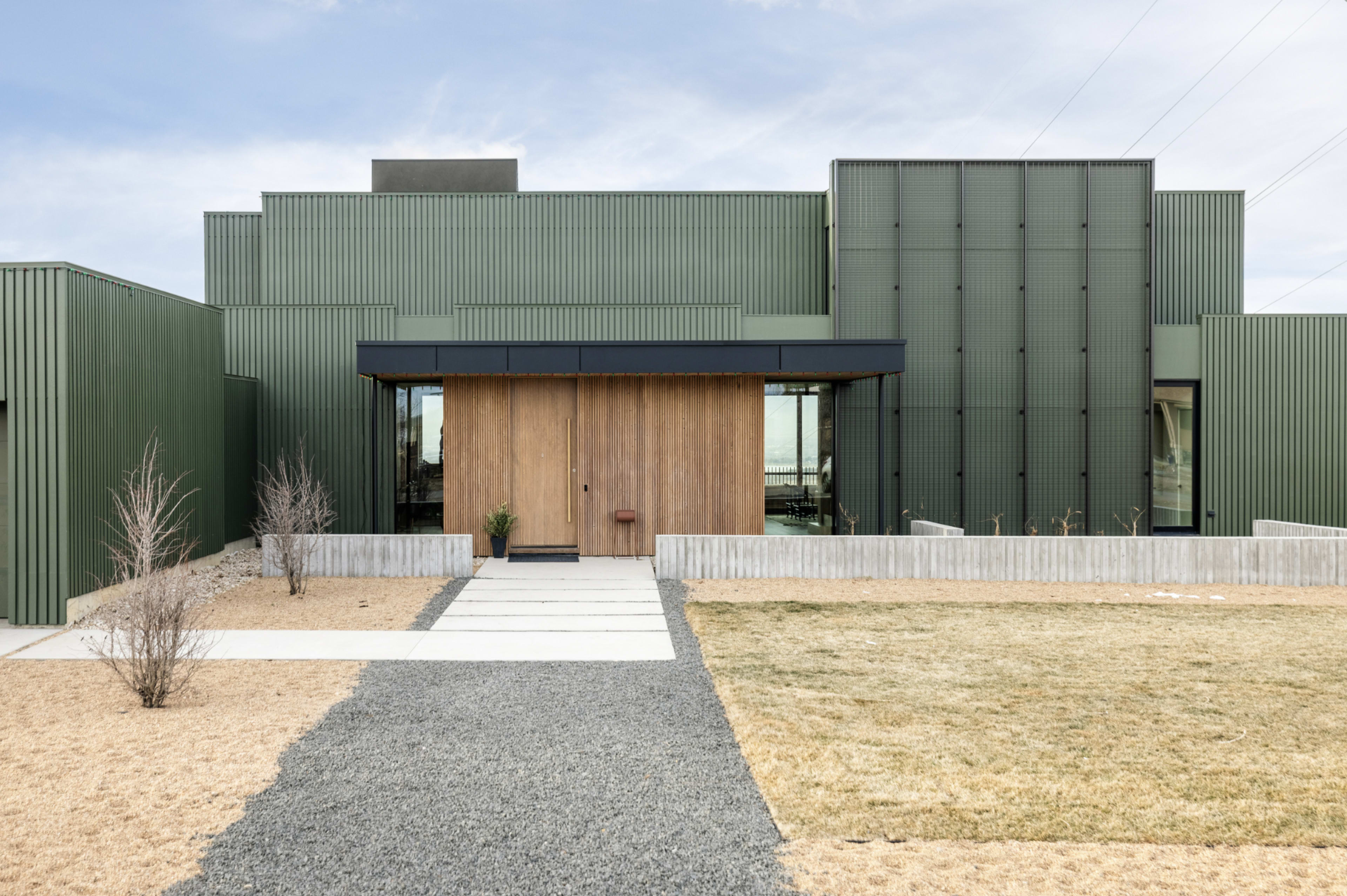 A modern house features a green metal exterior with a wooden front door, surrounded by a landscaped yard with gravel and grass.