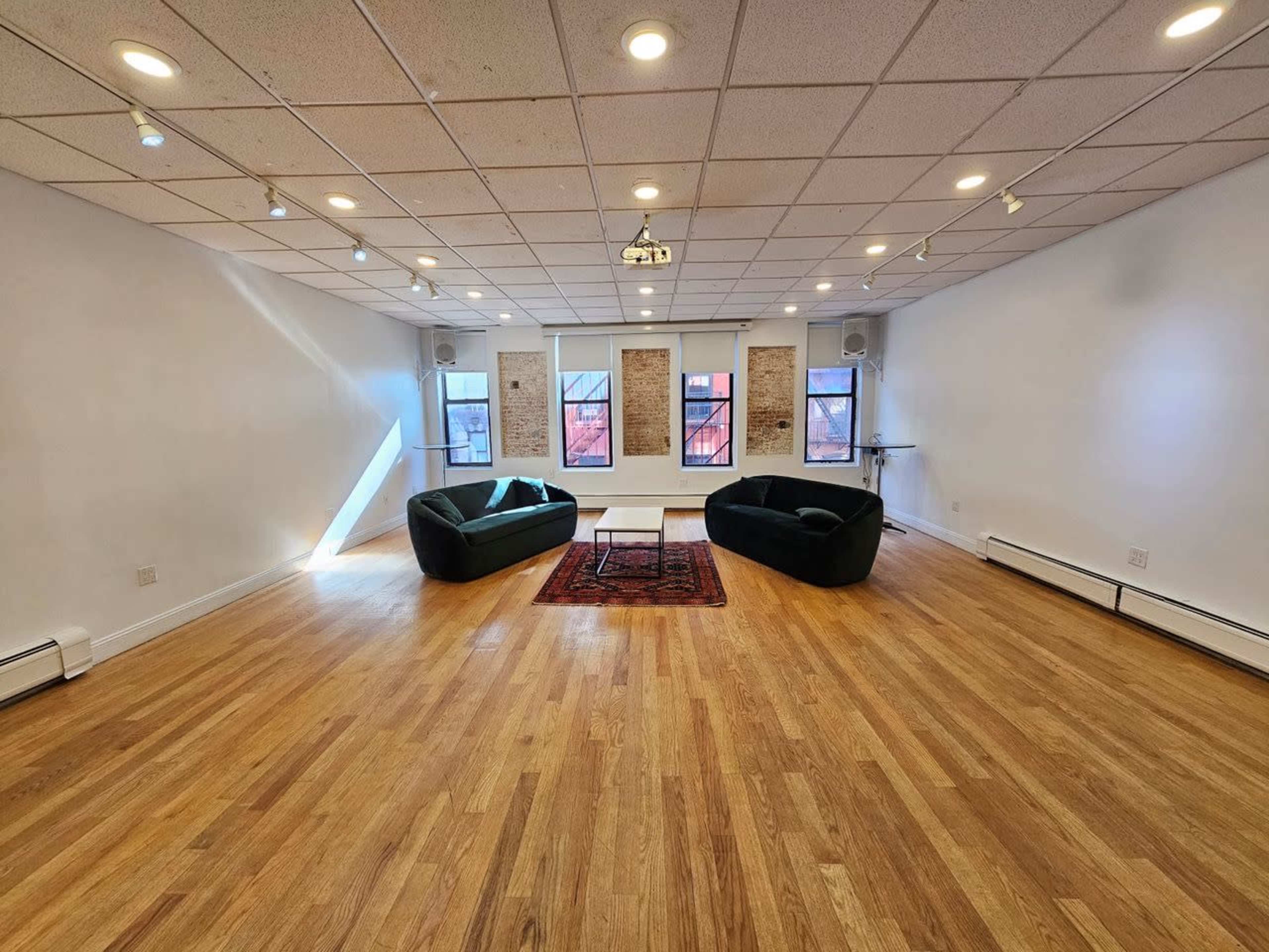 A spacious room with two dark green couches facing a central white table on a wooden floor, with several windows providing natural light.
