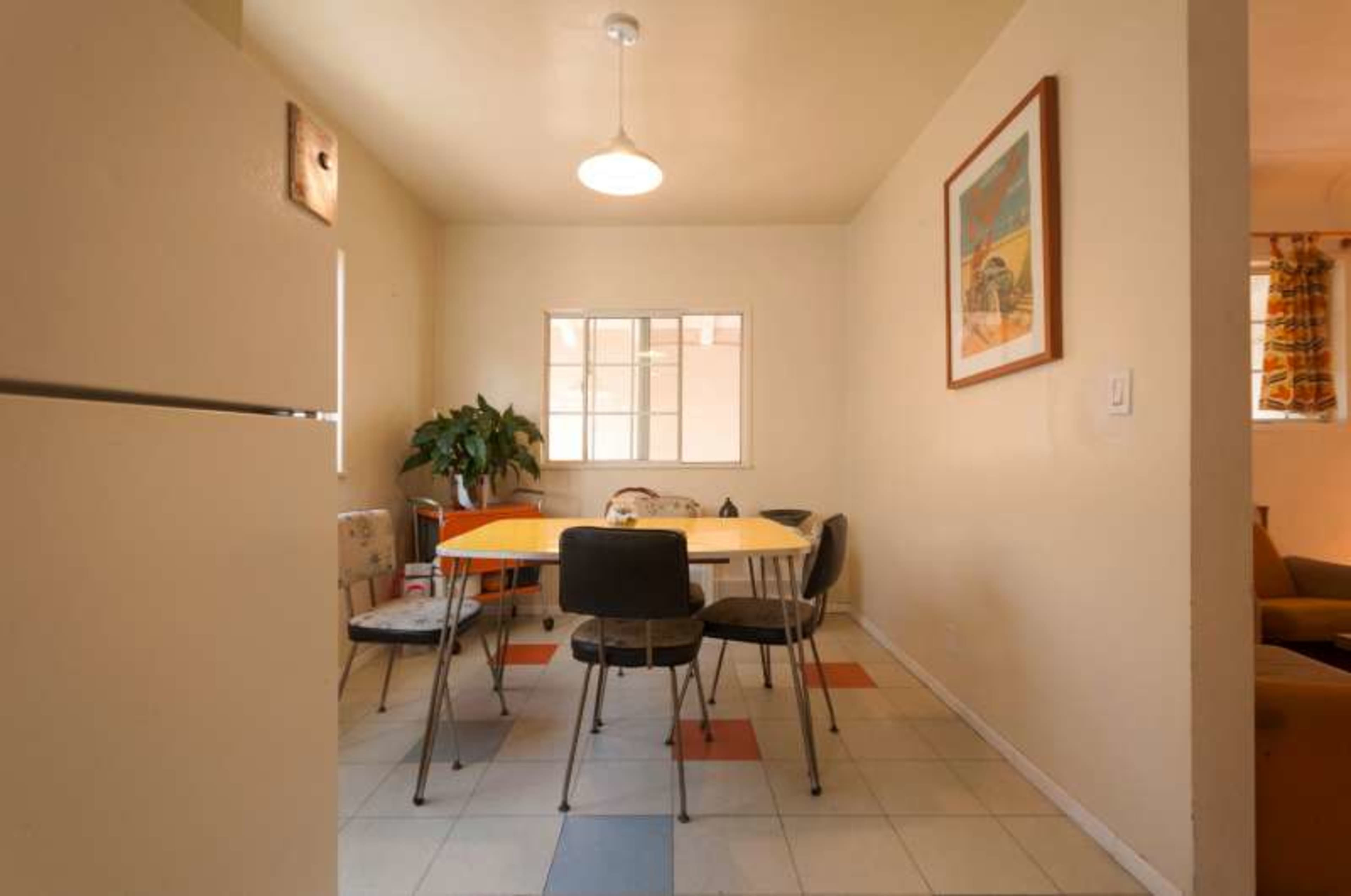 A small dining area features a table surrounded by chairs, with a window and a potted plant in the background.