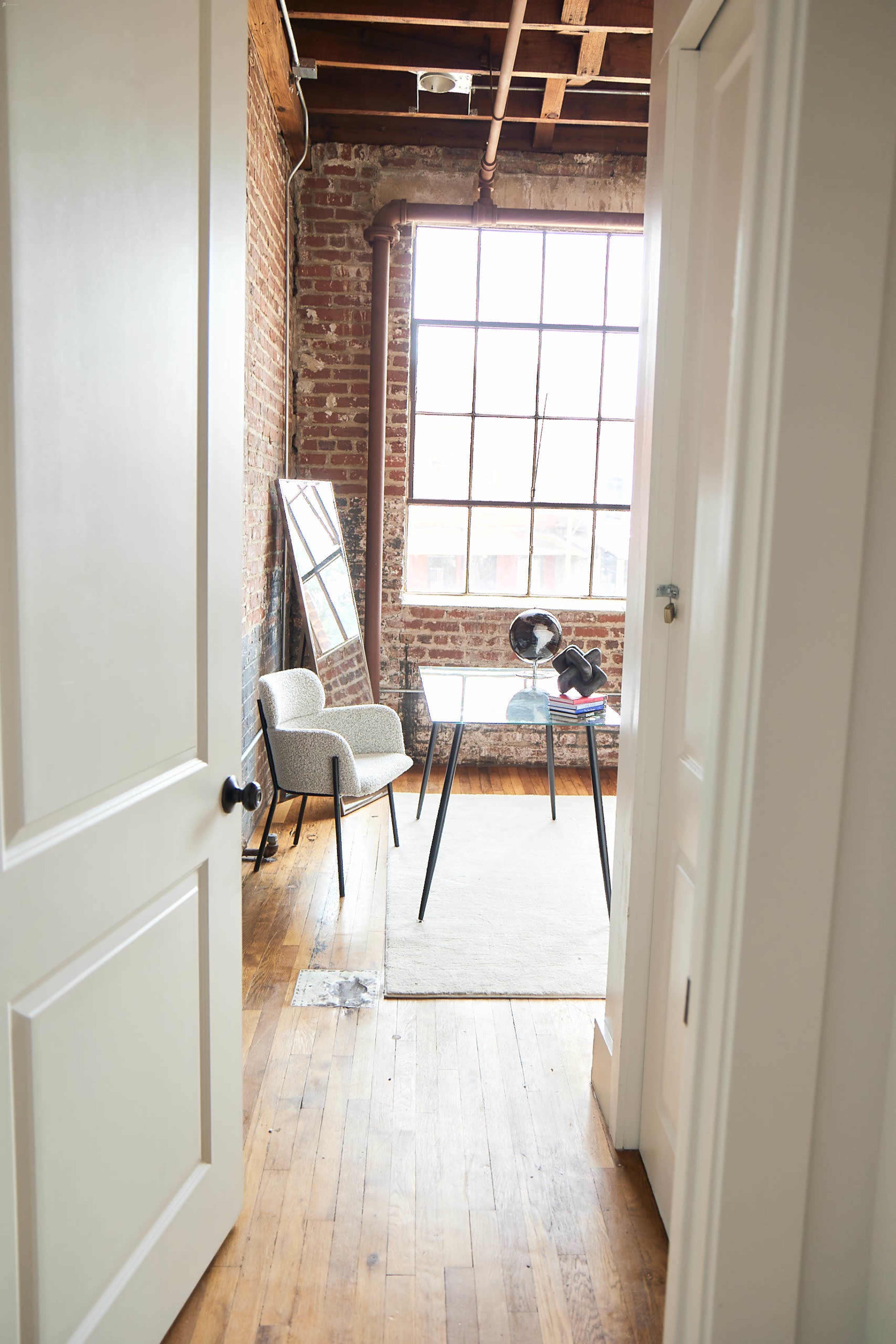 A well-lit room with a large window, brick walls, a small table, and a chair, seen through an open door.