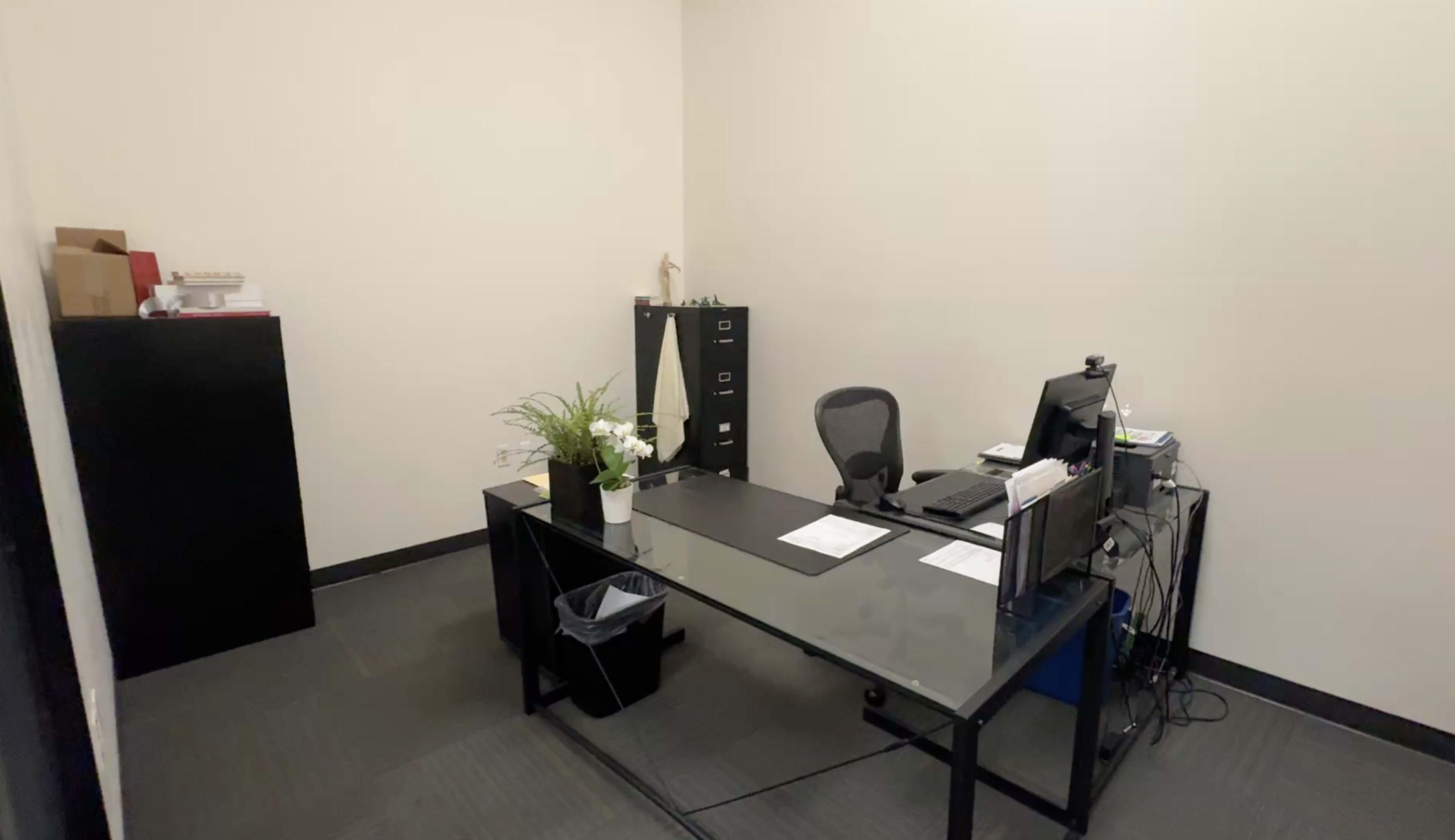 The image shows a small office space with a glass-top desk, computer, chairs, a filing cabinet, and a plant on the desk.