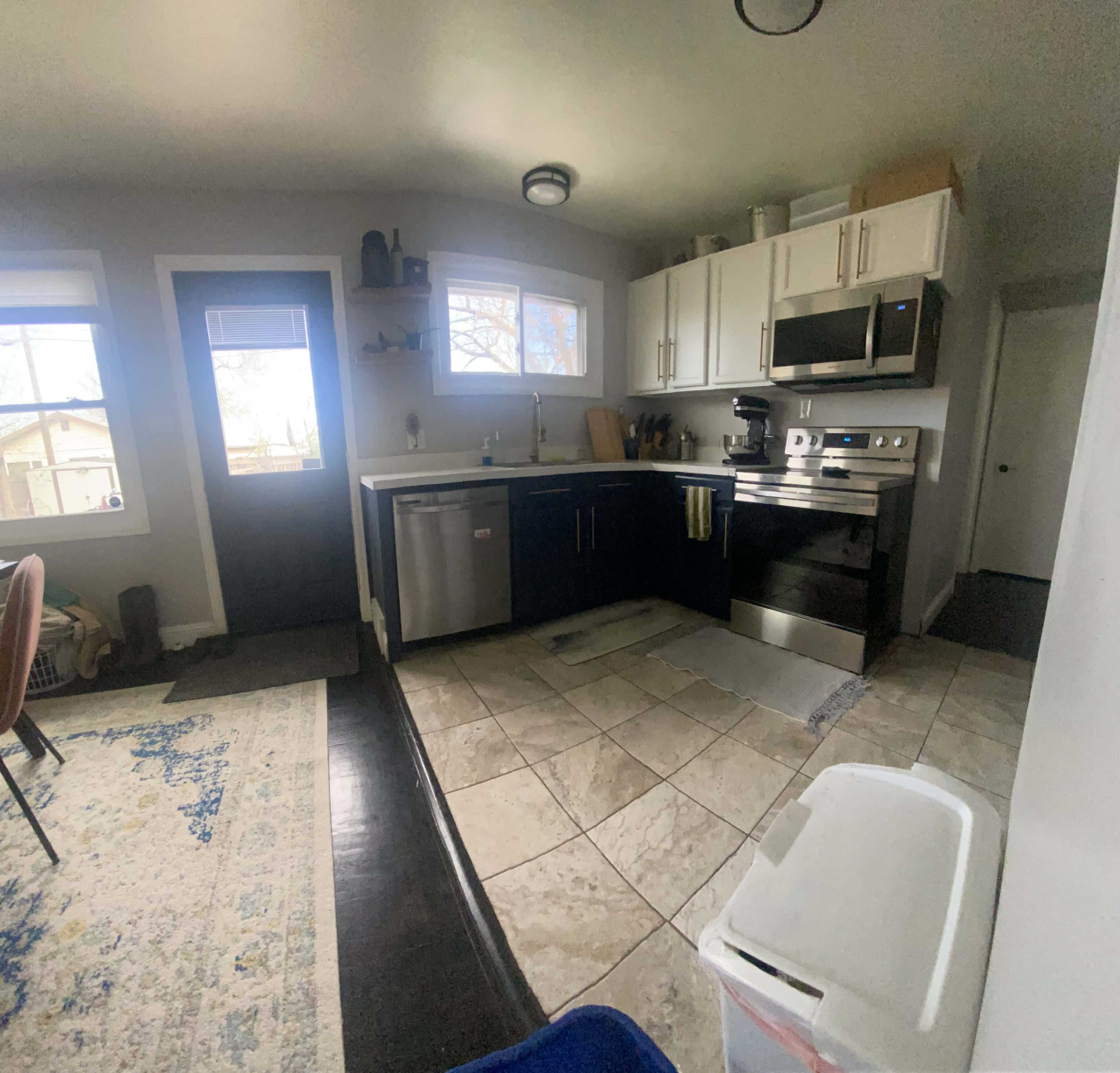 A modern kitchen with stainless steel appliances, a dark countertop, and tile flooring, connected to a dining area with a rug.