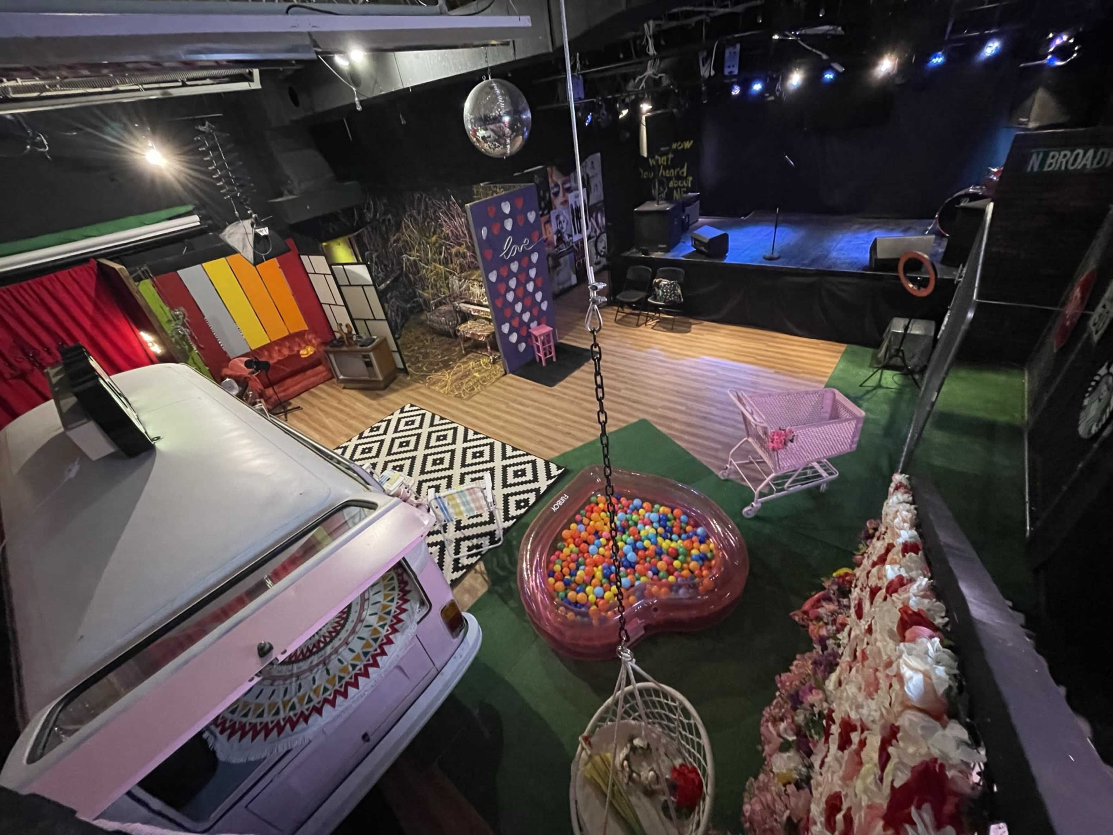 A colorful performance space featuring a retro van, a ball pit, hanging chairs, and a stage with blue lighting in the background.