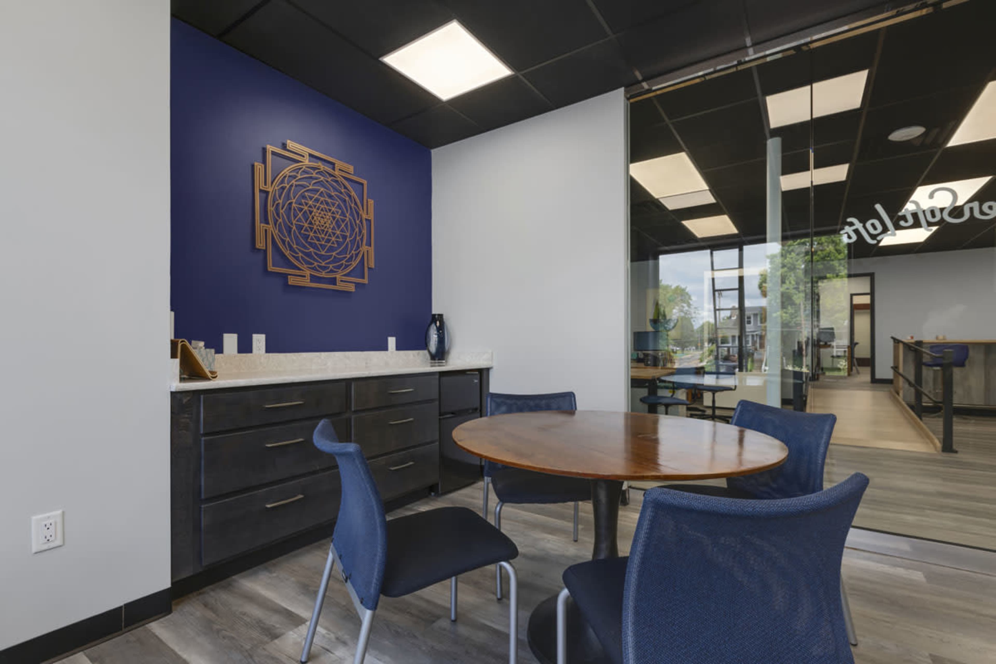 A small meeting area features a round wooden table surrounded by blue chairs, with a dark cabinet and a decorative wall art piece in the background.