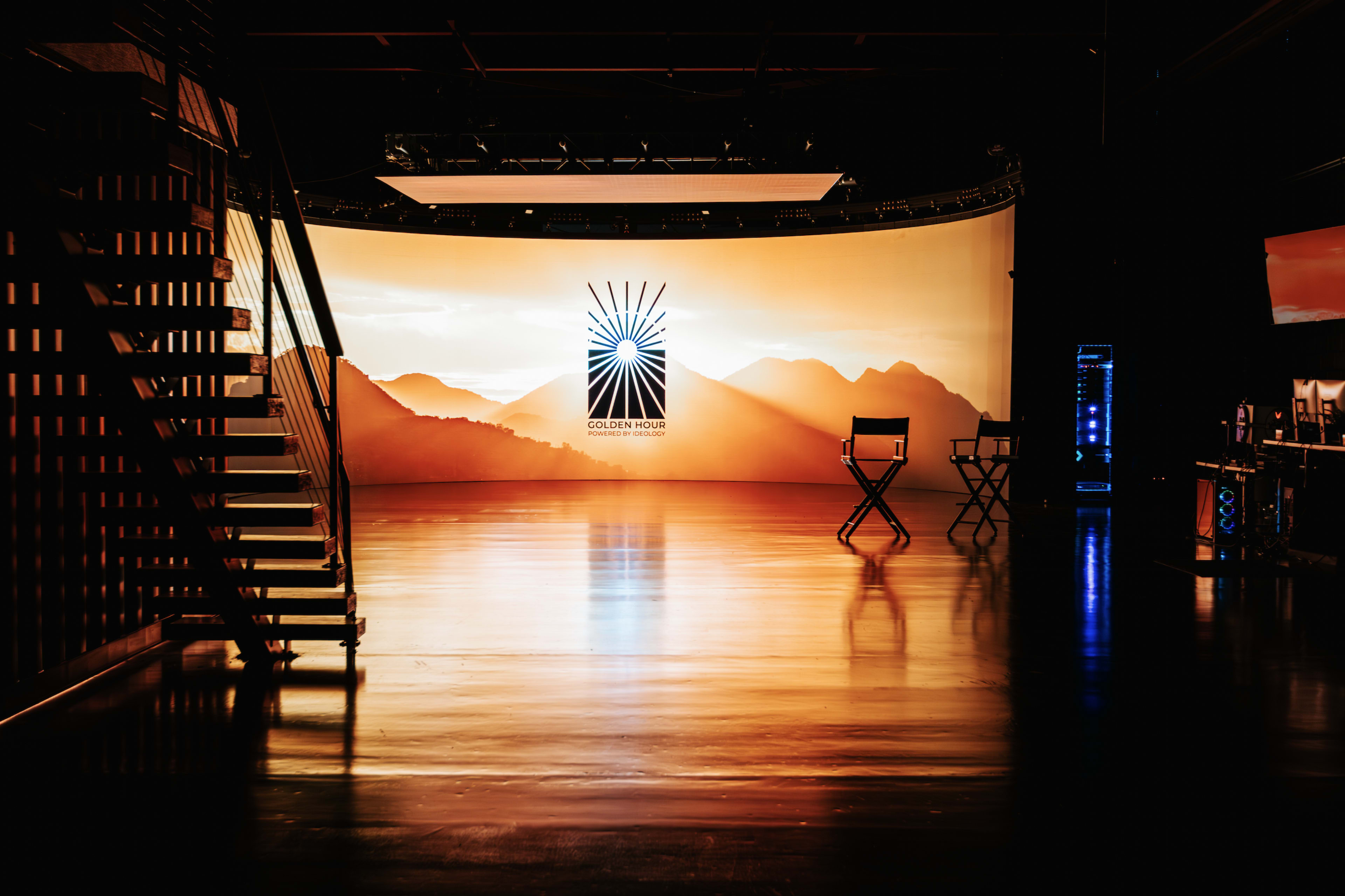 A film or television studio features a large backdrop with a mountain silhouette and a logo, along with a director's chair on a shiny floor.