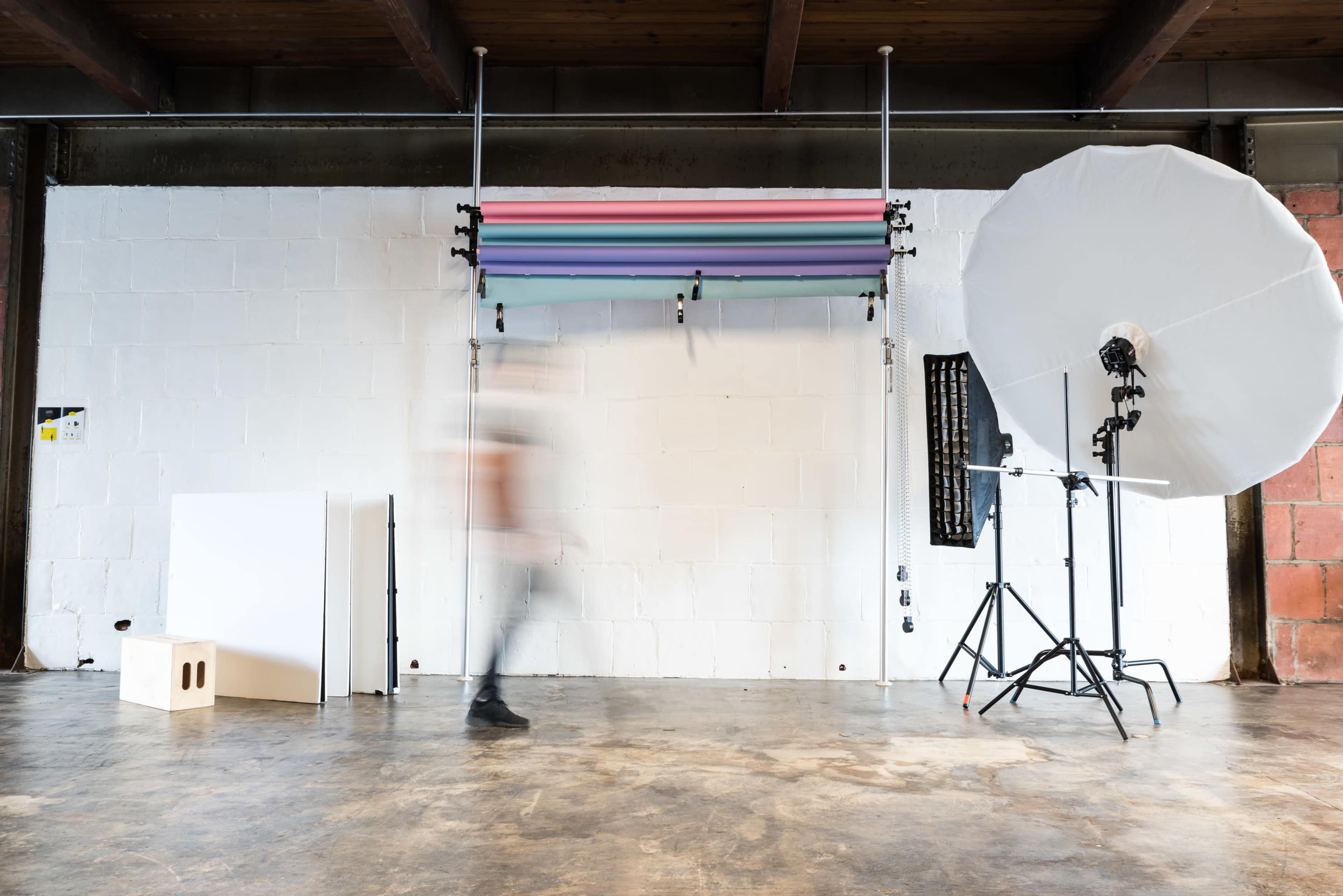 A blurred figure walks past a photography setup featuring backdrop rolls and studio lighting in an industrial space.