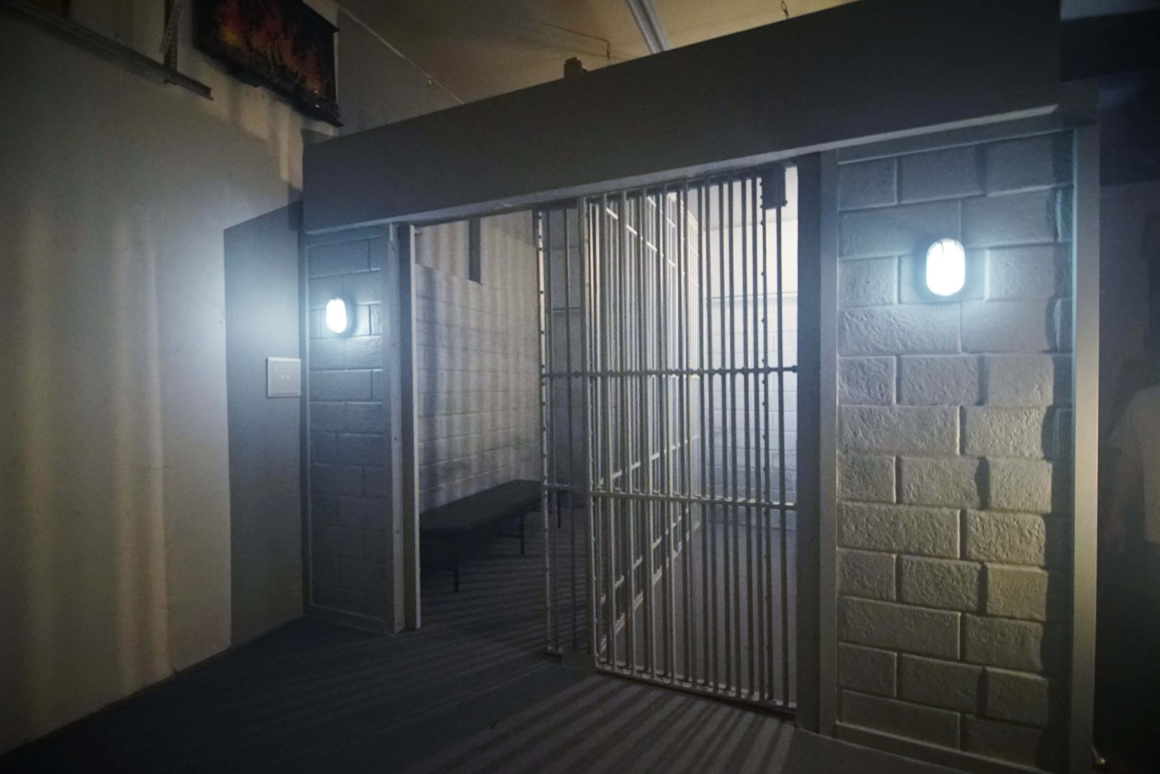 A dimly lit corridor with a barred entrance leading to a cell, flanked by two wall-mounted lights.