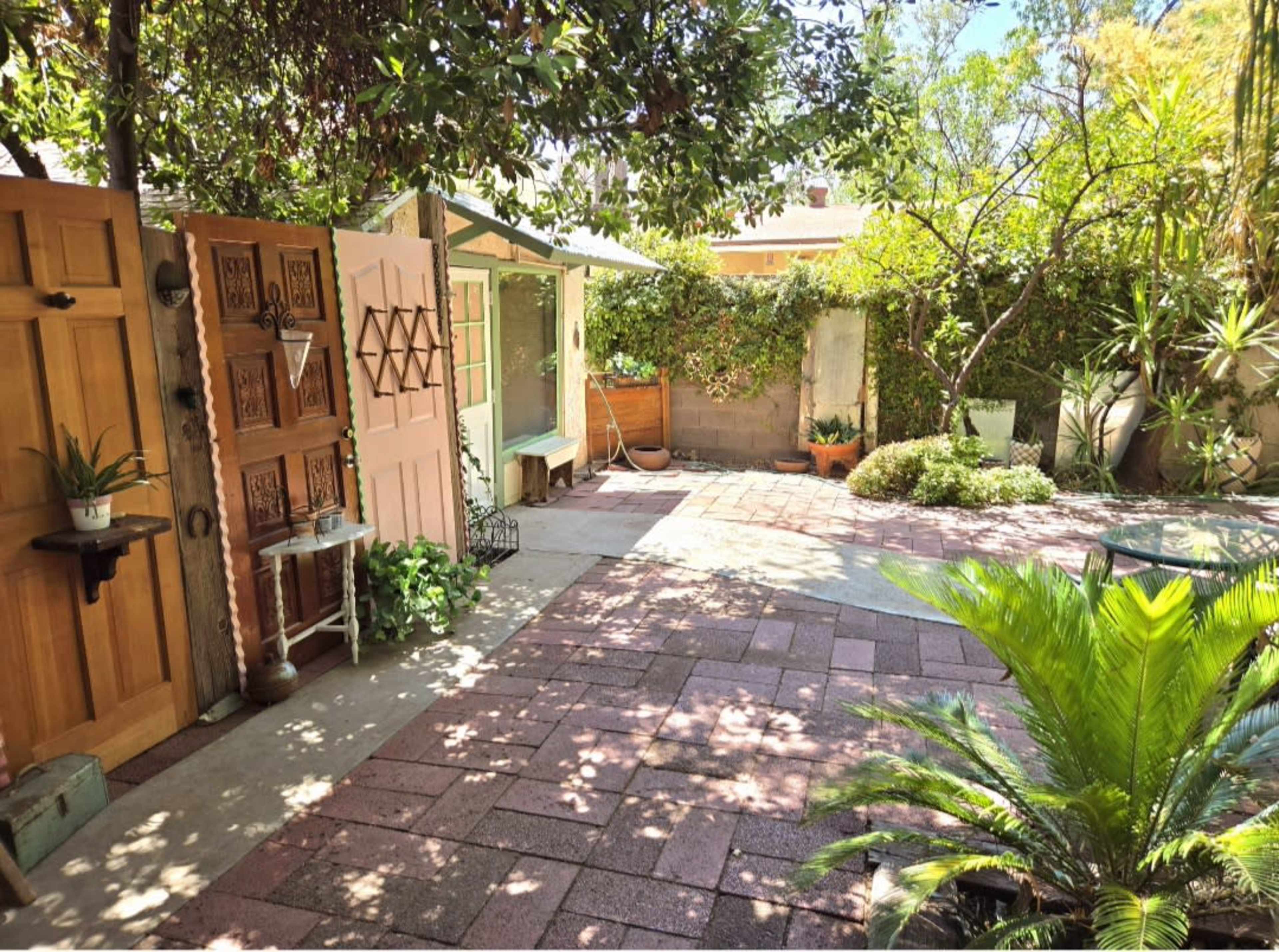 The image shows a backyard garden with stone pavement, potted plants, a wooden shed, and decorative wooden doors.