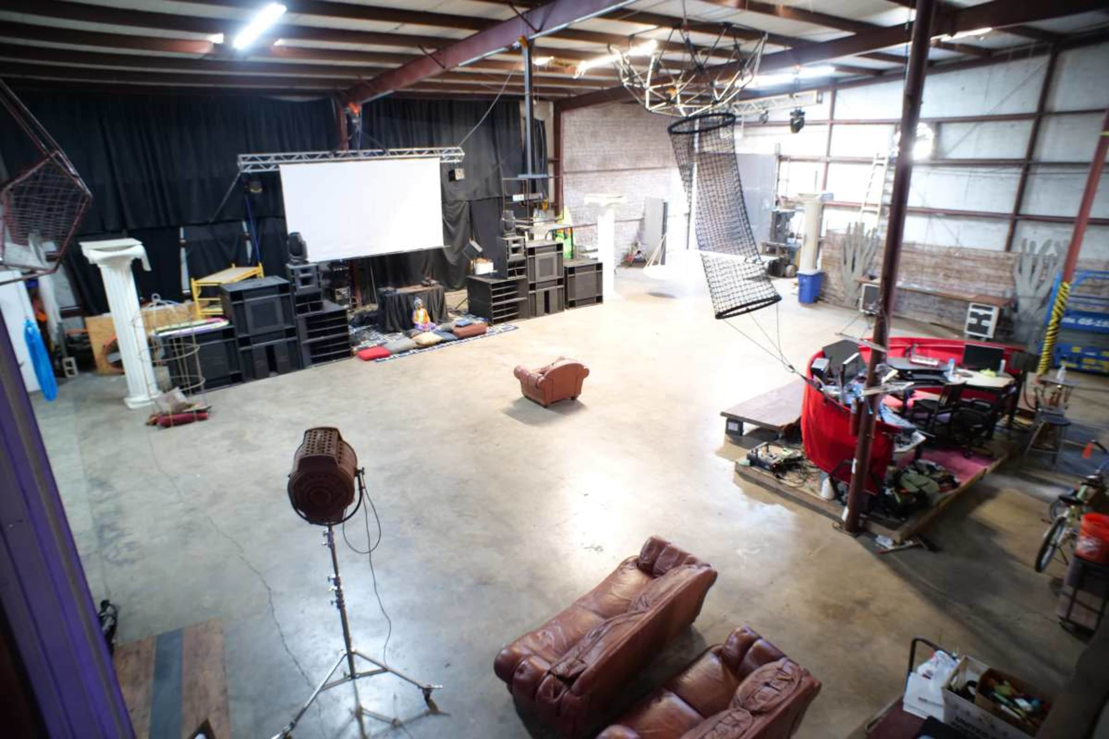 The image shows a spacious venue with a large screen, sound equipment, and seating arrangements, including two brown leather couches in the center.