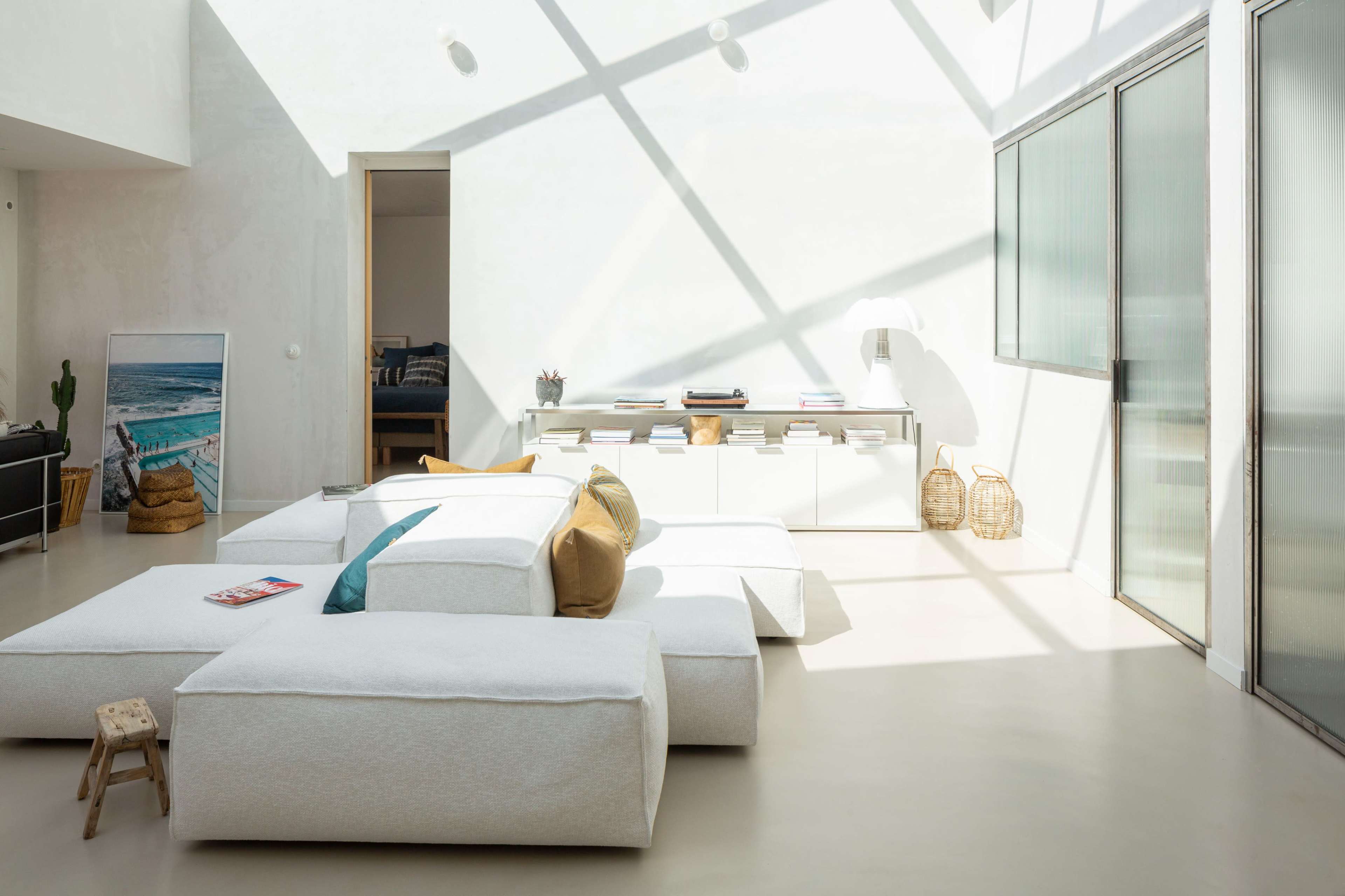 The image shows a bright, minimalist living room featuring large cushioned seating, a low table, and a window with natural light casting geometric shadows on the floor.