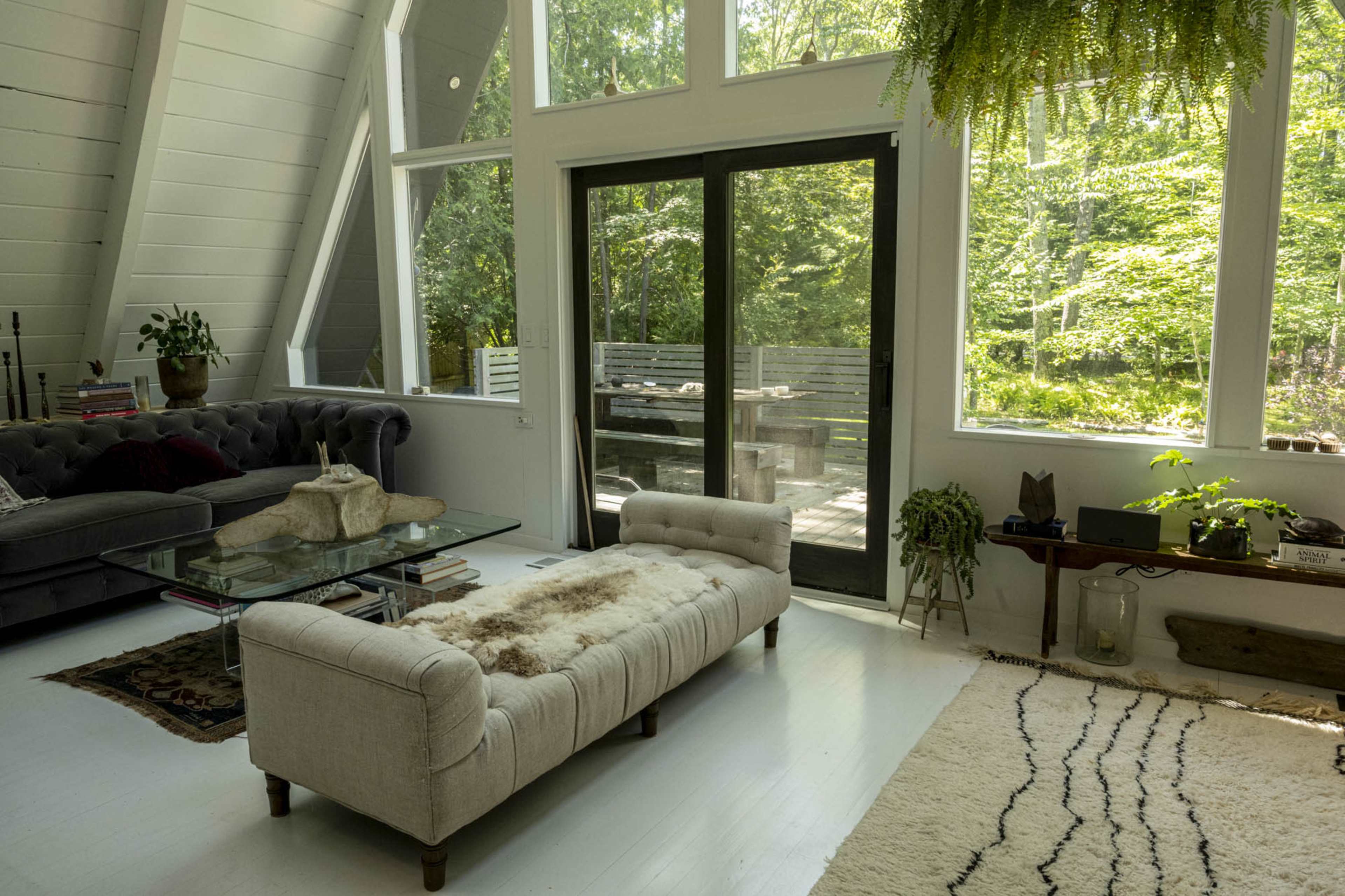 A modern living room features a tufted sofa, a glass coffee table, and large windows that overlook a wooded area.