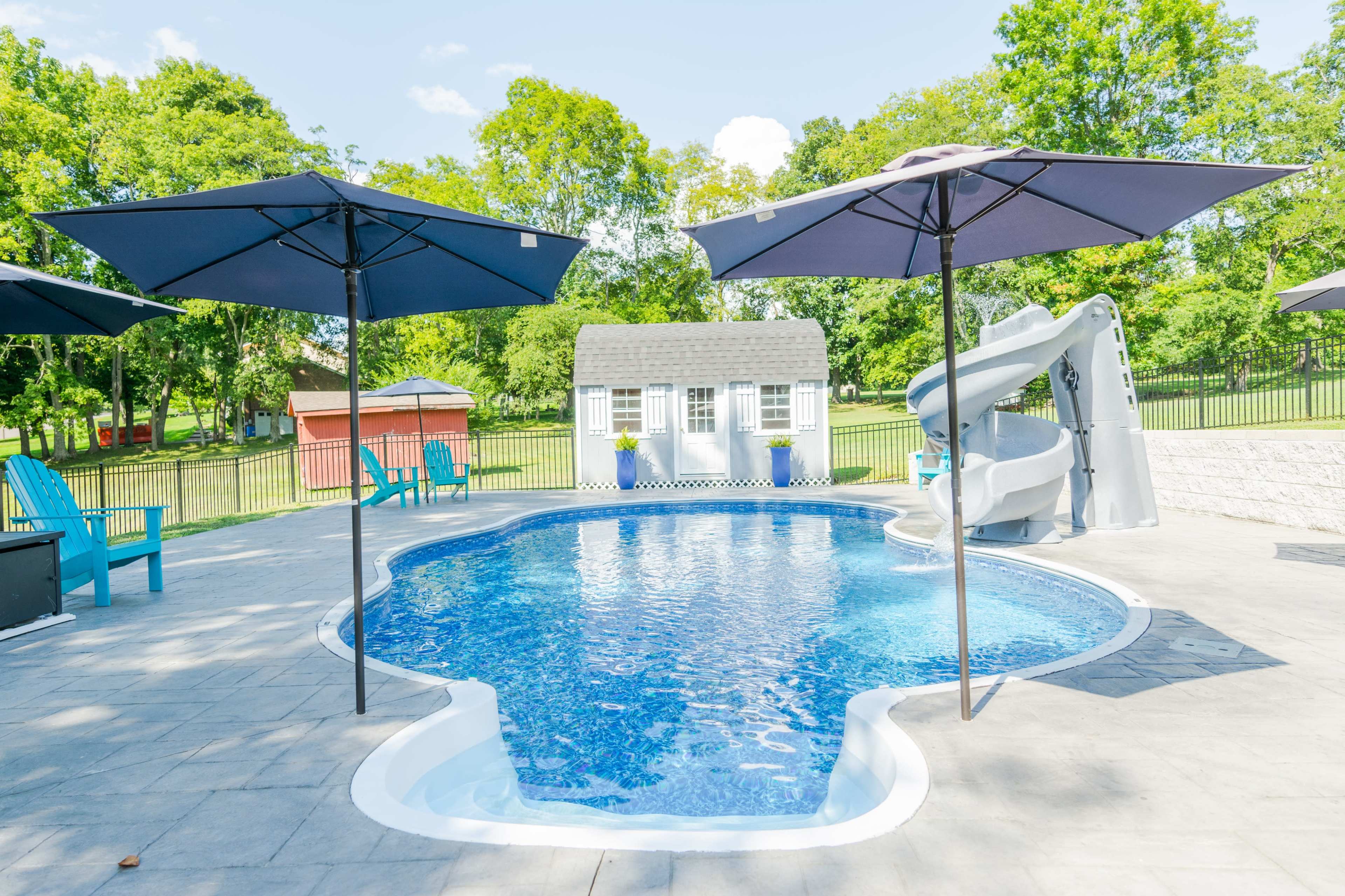 A swimming pool surrounded by lounge chairs and umbrellas, featuring a water slide and a small building in the background.