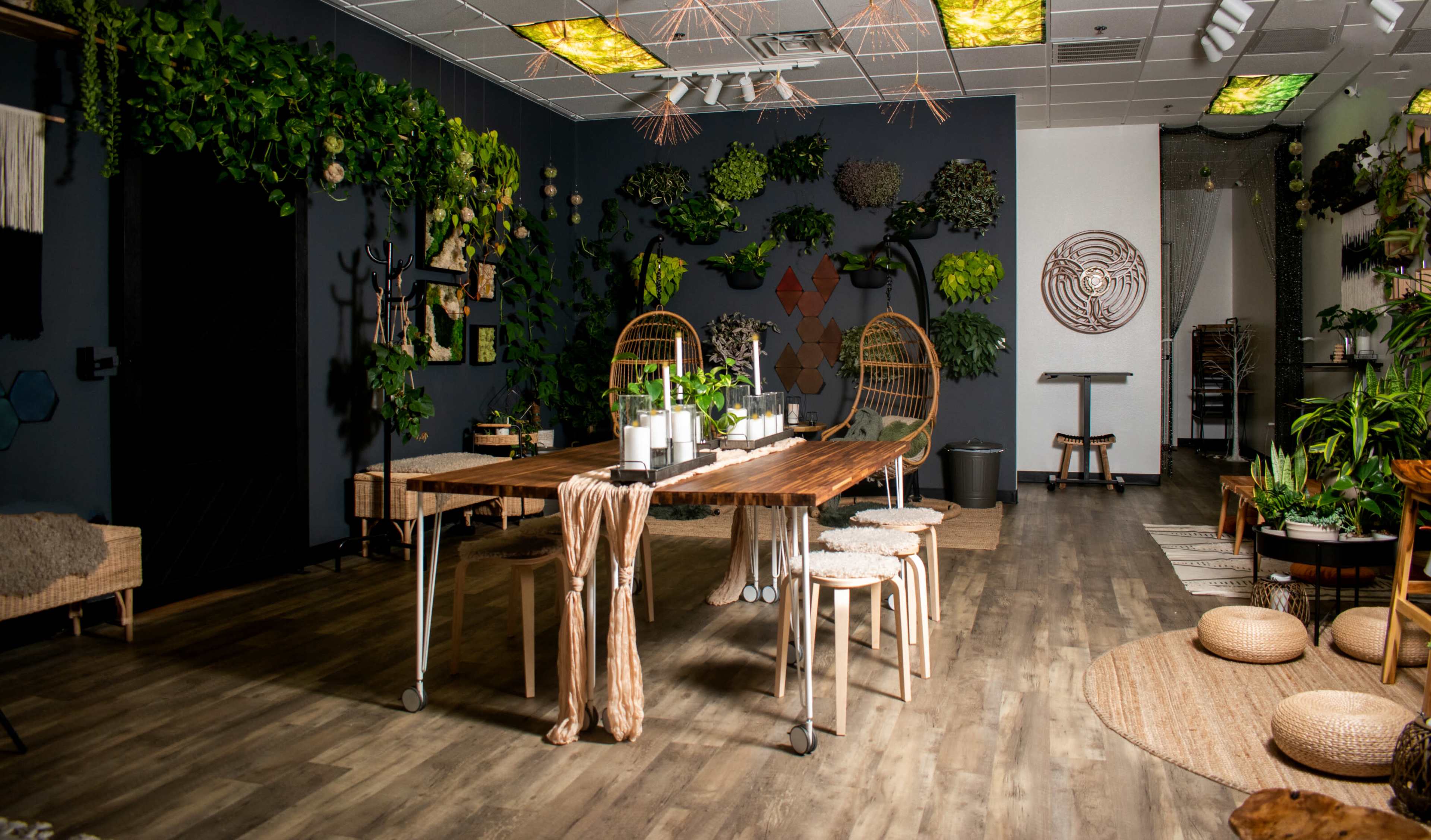 A brightly lit indoor space features a long wooden table surrounded by stools, with a backdrop of greenery and decorative wall art.