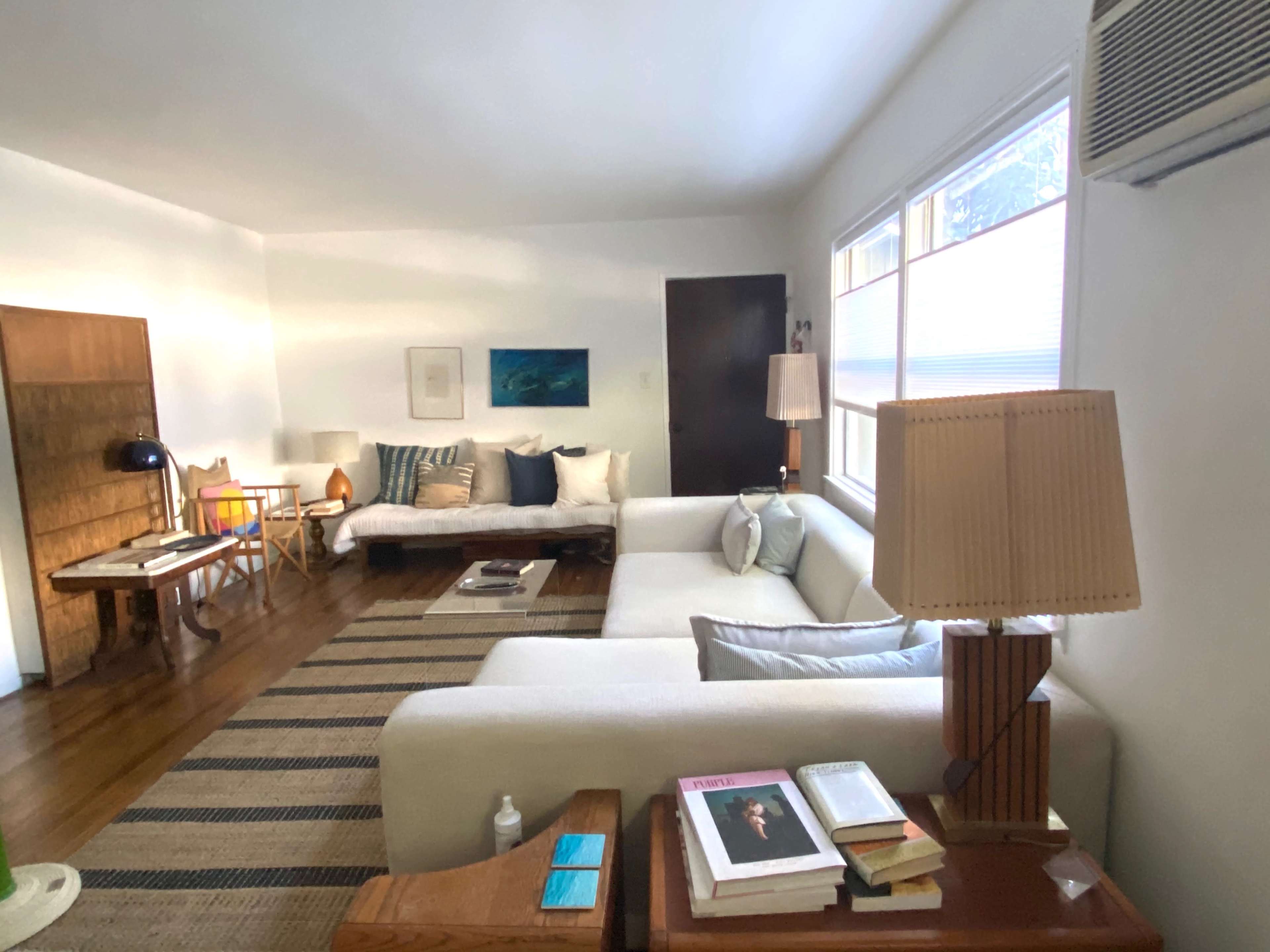 The image shows a bright living room featuring a white sectional sofa, a wooden coffee table with books, and a large window letting in natural light.