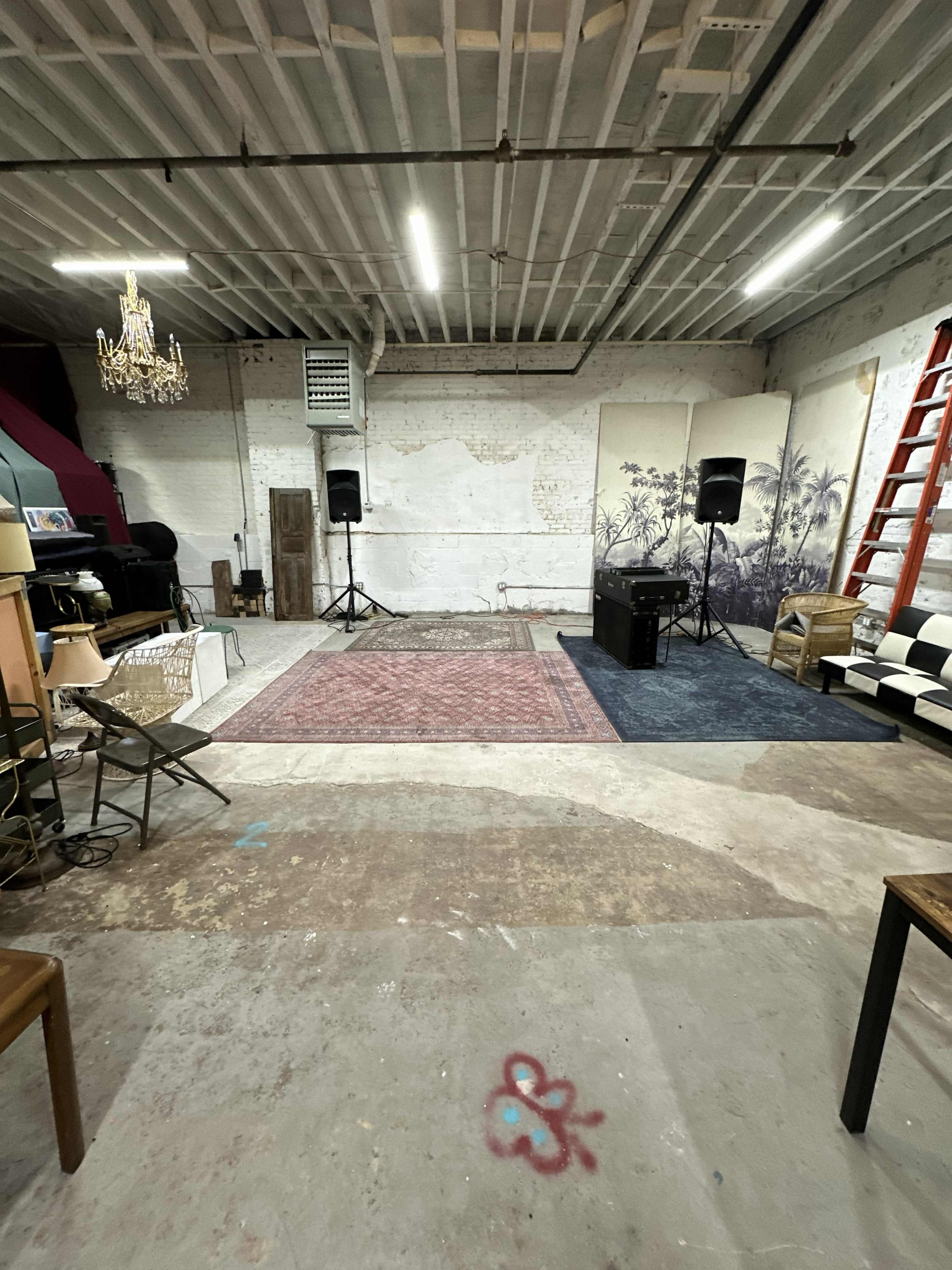 The image shows an empty, industrial-style room with exposed brick walls, two speakers set up on a wooden floor, and various furniture items including tables and chairs around the space.