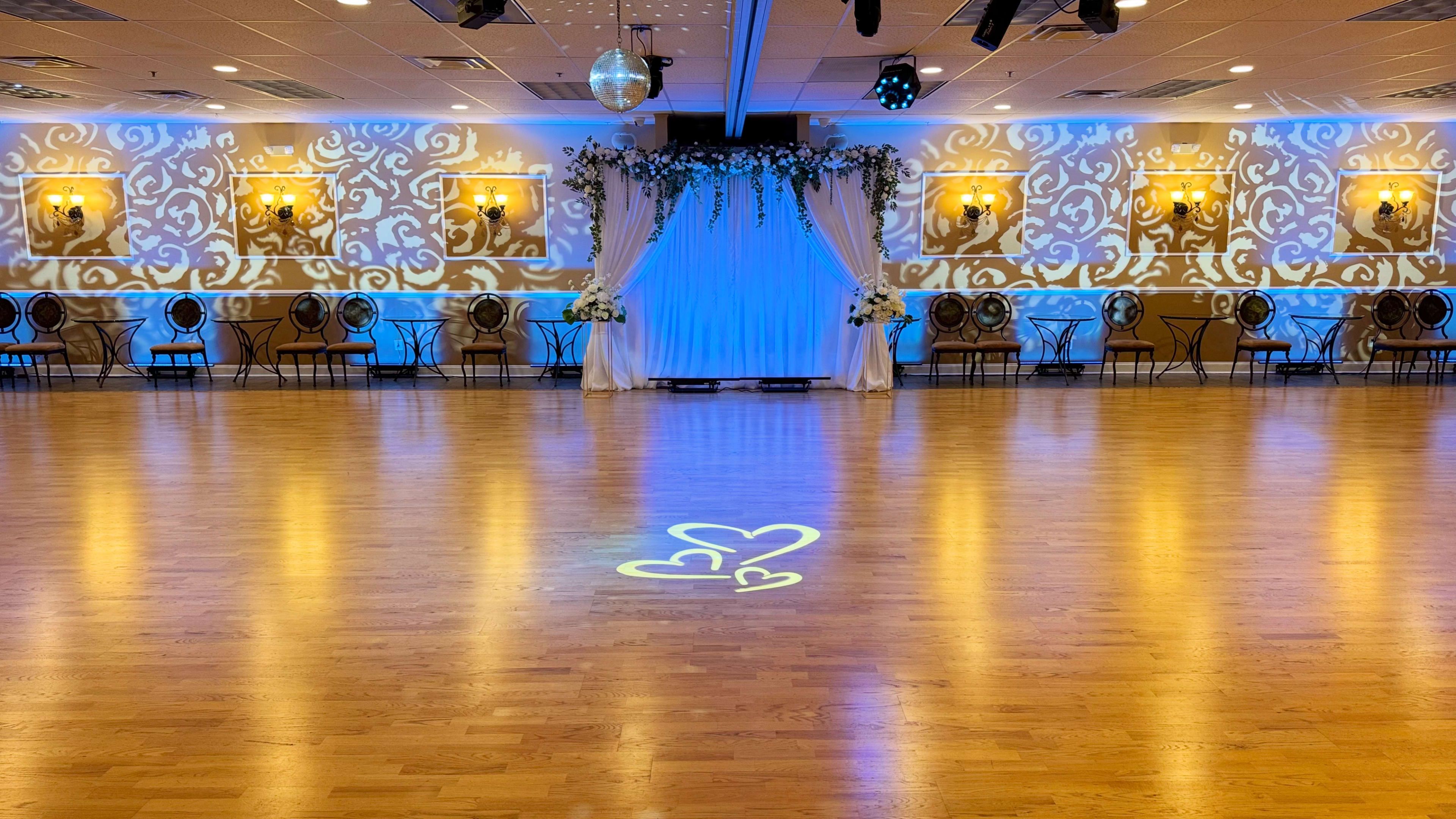 The image shows an elegantly decorated dance hall with wooden flooring, a floral backdrop, and soft blue lighting reflecting on the walls.