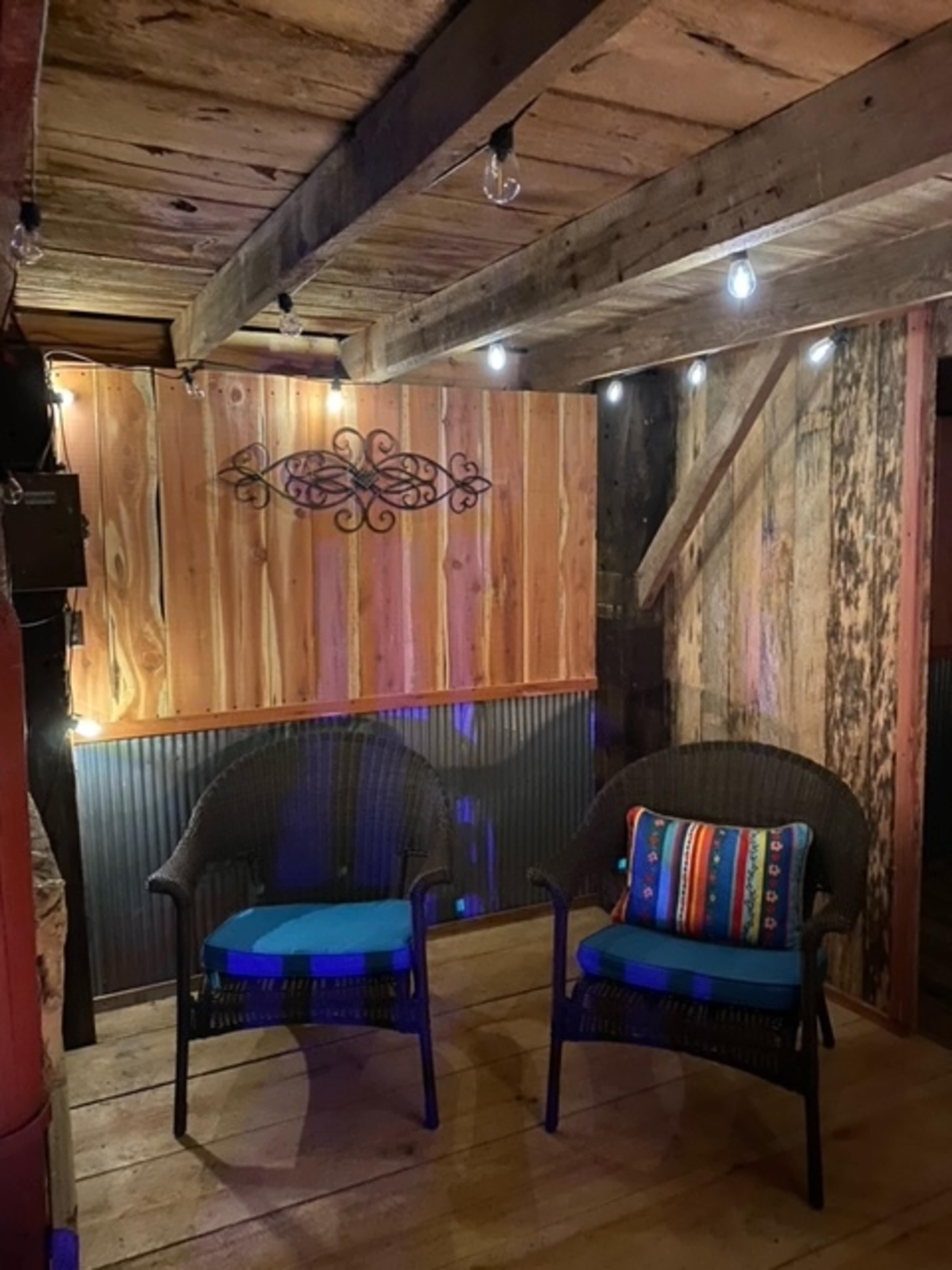 The image shows a rustic interior featuring two black wicker chairs with blue cushions, a wooden wall with decorative artwork, and string lights overhead.