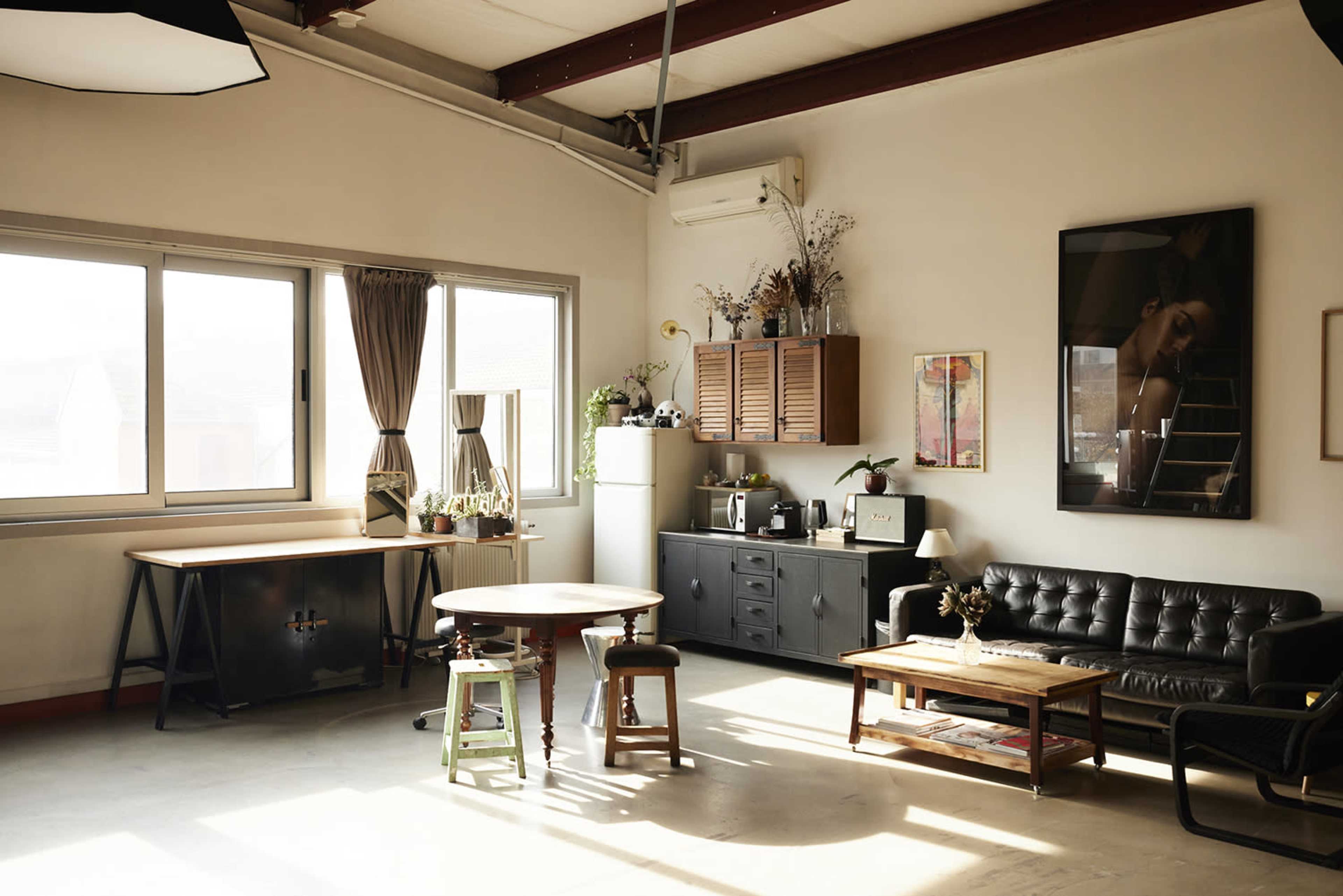 The image depicts a well-lit, minimalist studio apartment with a black leather sofa, a wooden table, and various decorative plants.