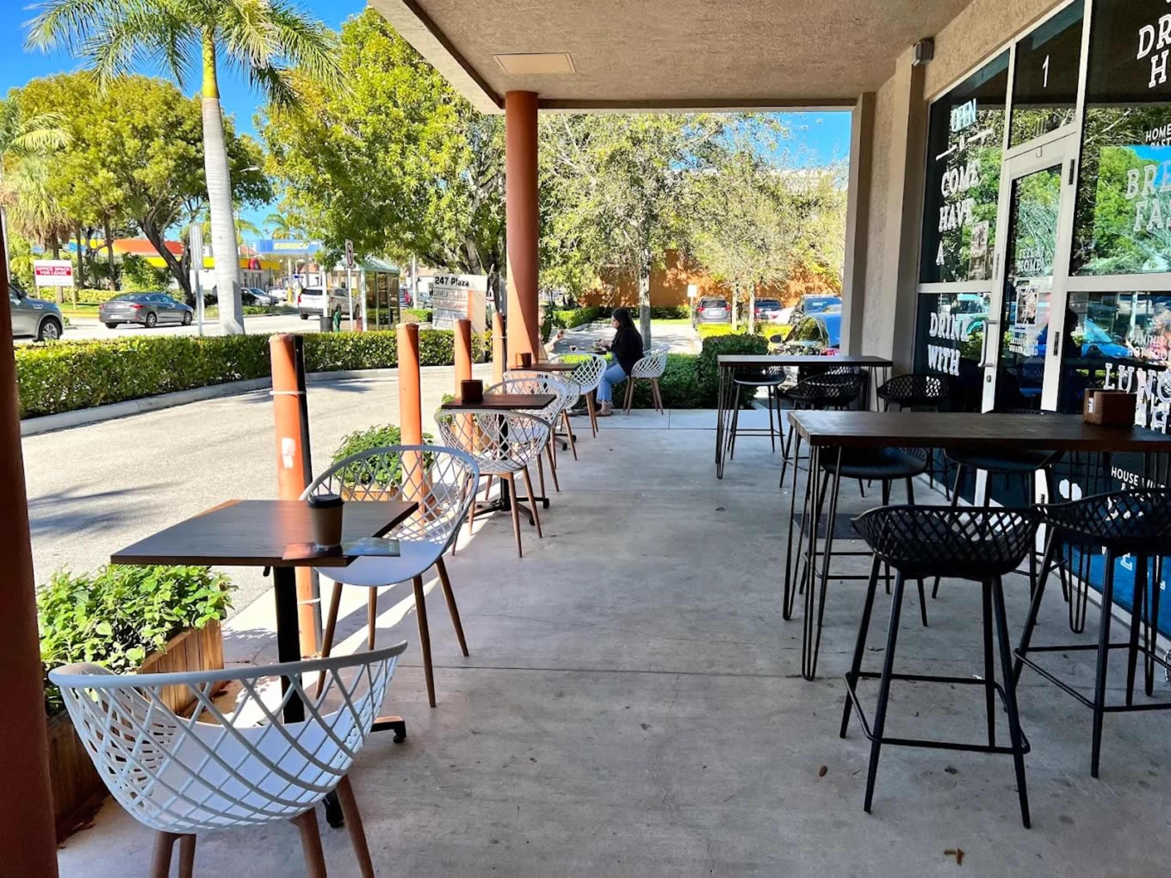 The image shows a patio area of a café with tables and chairs situated outside under a canopy, surrounded by greenery and parked cars in the background.