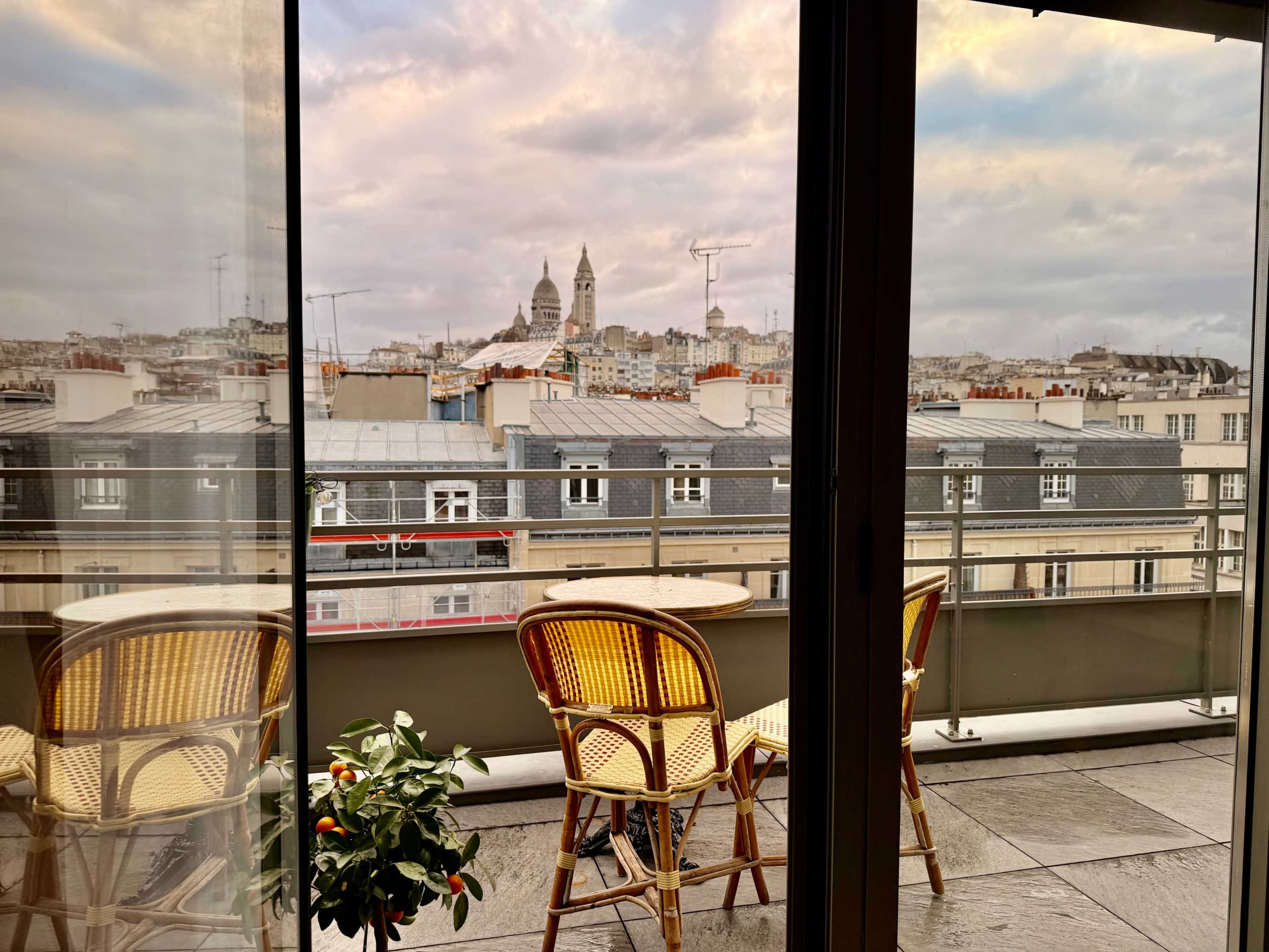 The image shows a balcony with two wicker chairs facing a cityscape featuring prominent buildings under a cloudy sky.