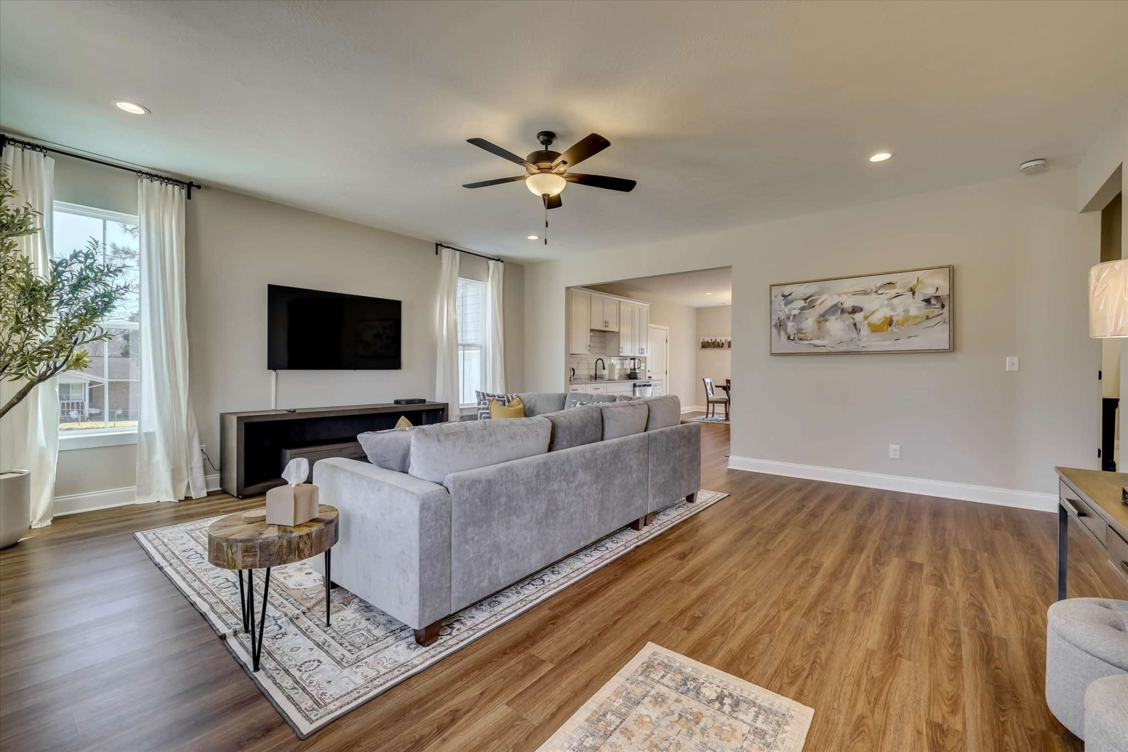 The image shows a modern living room featuring a gray sofa, a wooden coffee table, a flat-screen TV mounted on the wall, and large windows with sheer curtains, leading to an open kitchen area.