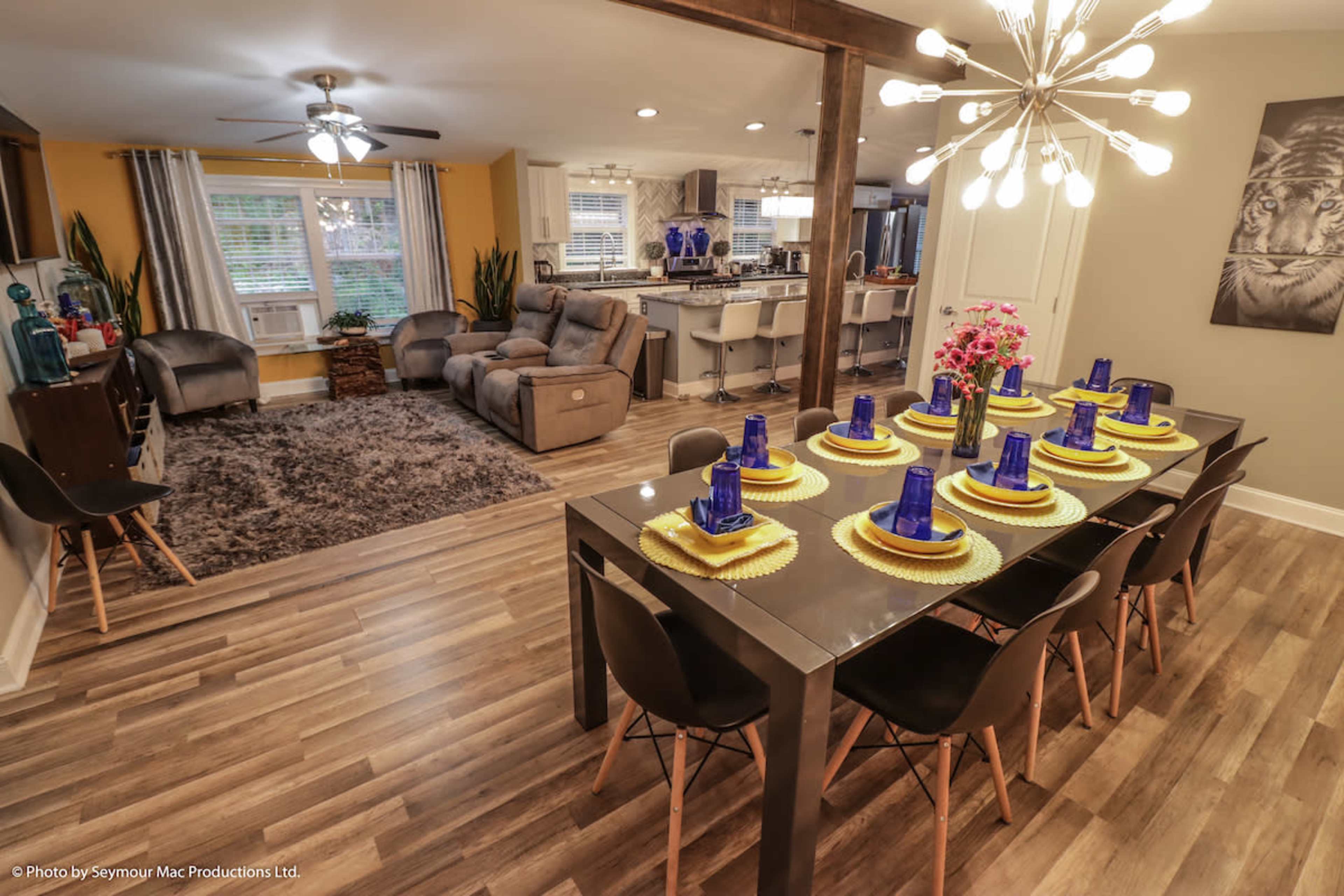 A dining area with a set table featuring blue cups and yellow plates, adjacent to a living area with a sofa and plants.