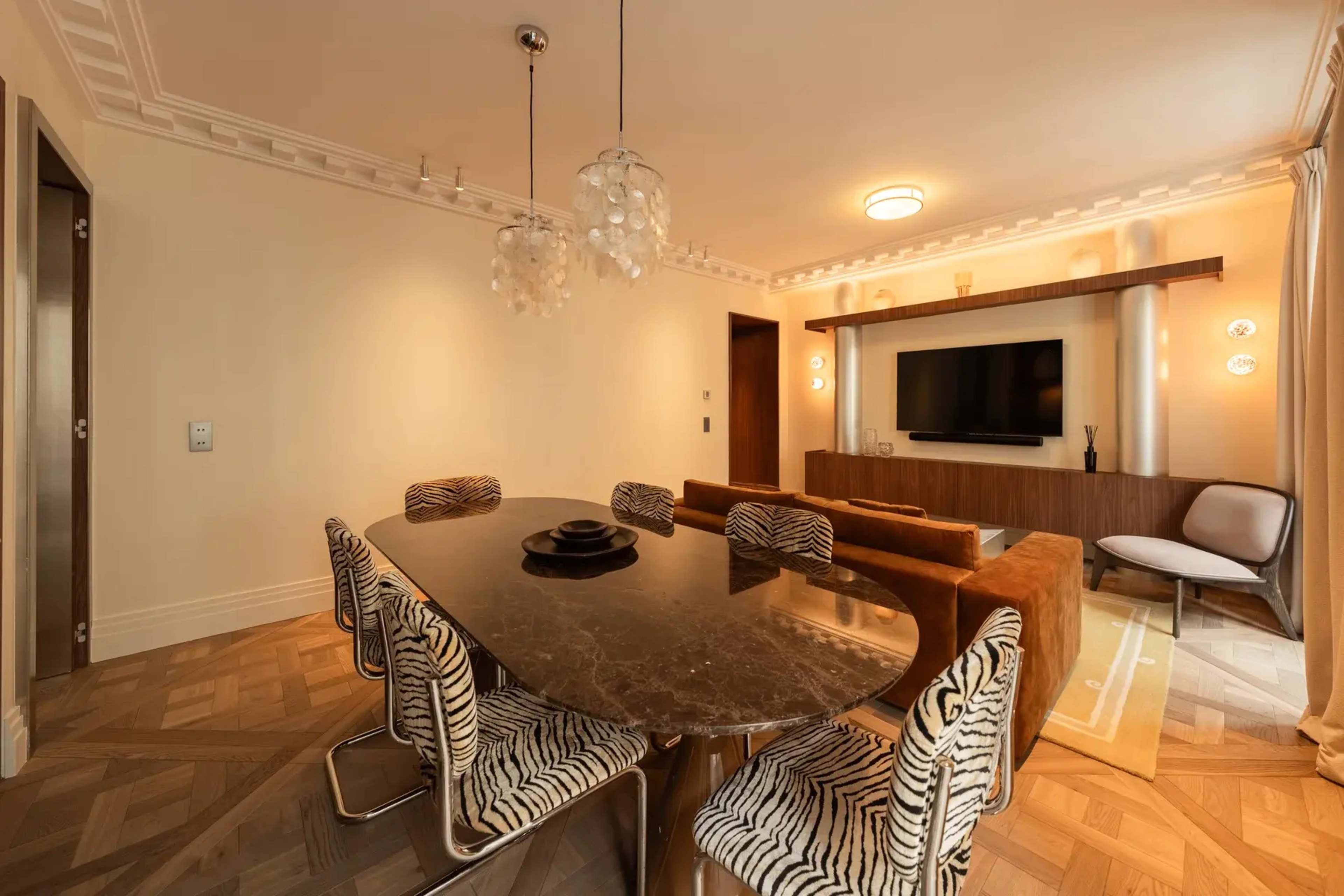 A spacious dining room features a large marble table surrounded by striped chairs, with a cozy seating area and a wall-mounted television in the background.