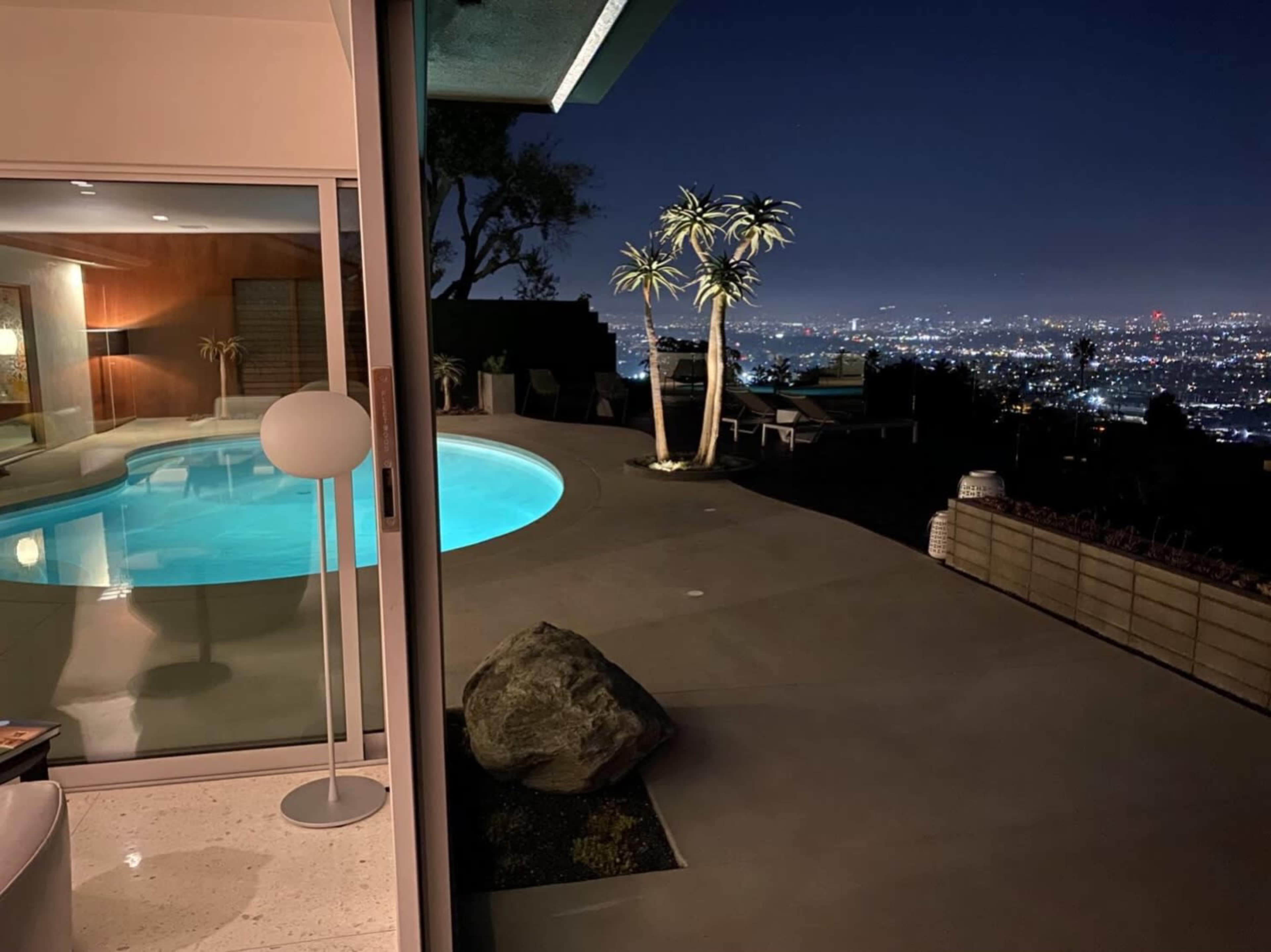 A modern home with a swimming pool overlooks a city skyline at night, featuring outdoor seating and a landscape with palm trees.