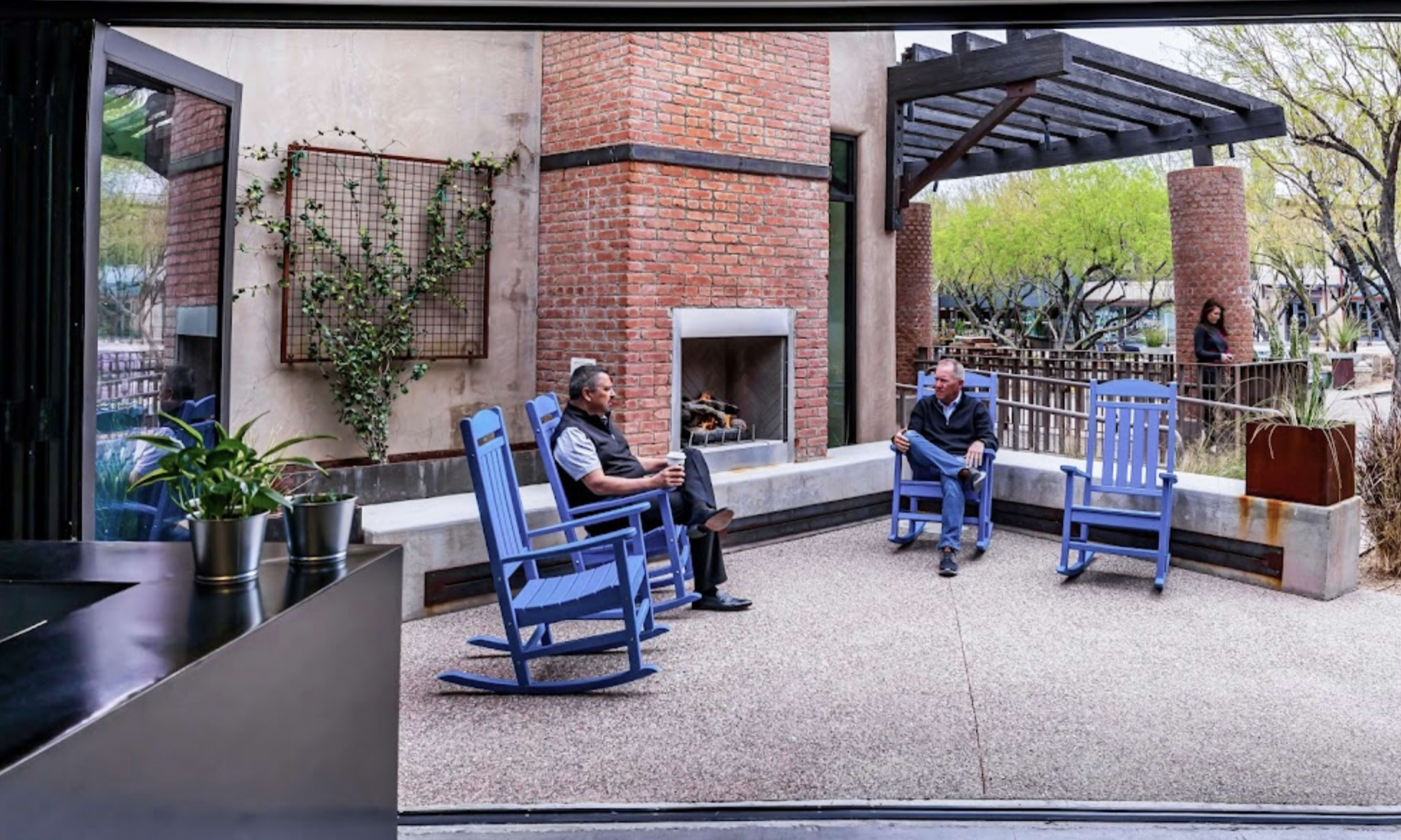 Two men sit in rocking chairs on a patio, with a brick fireplace and greenery in the background.
