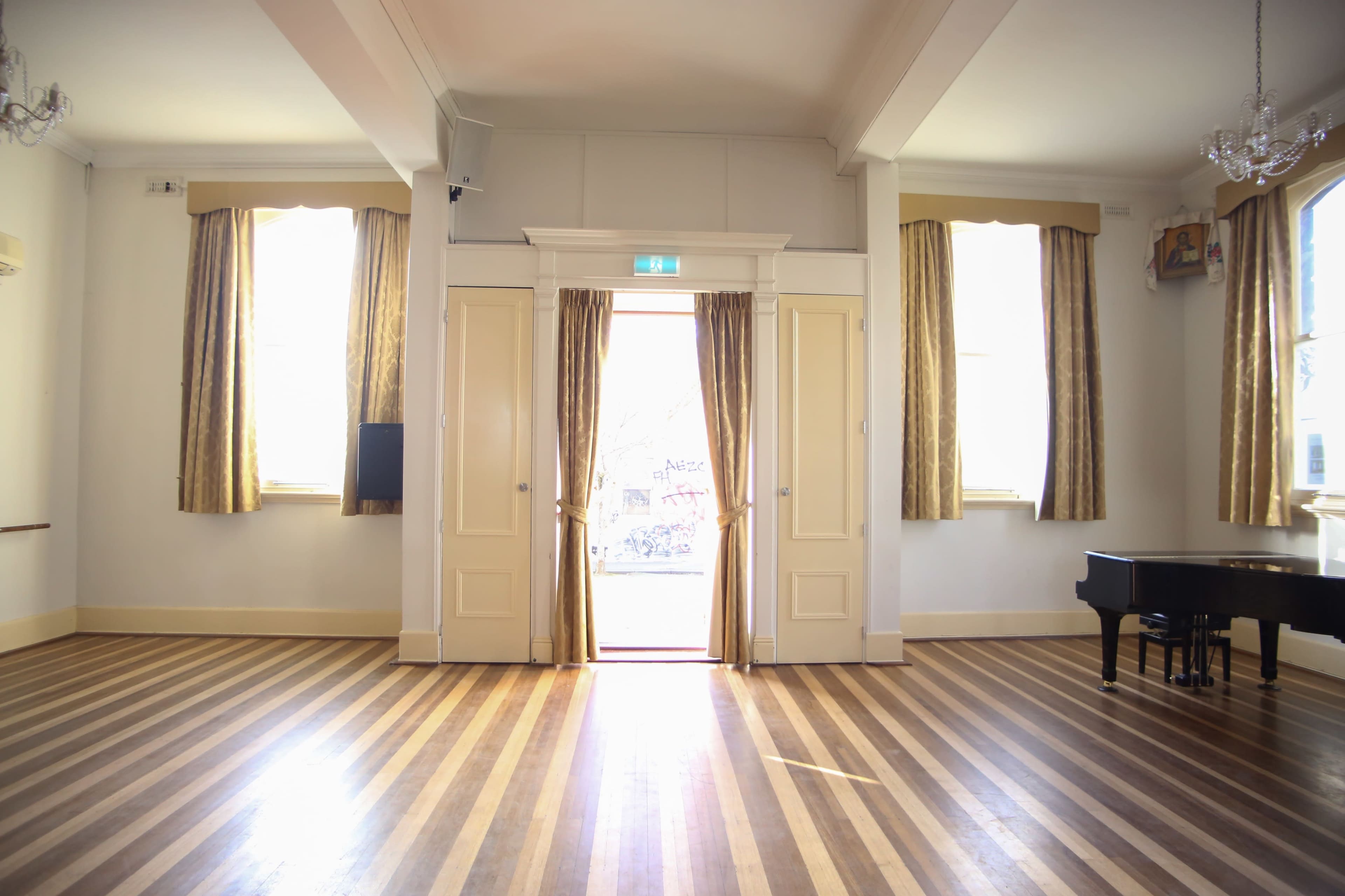 A spacious room with wooden flooring, large windows adorned with curtains, and a grand piano positioned against one wall.