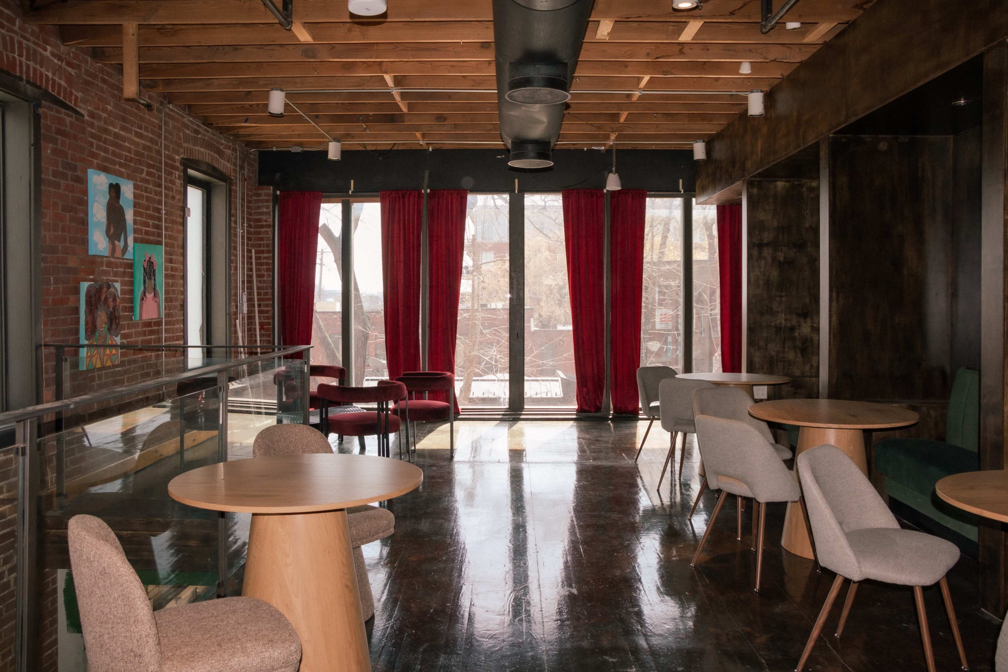 The image shows a modern interior space with wooden beams, large windows draped with red curtains, and several round tables surrounded by chairs.