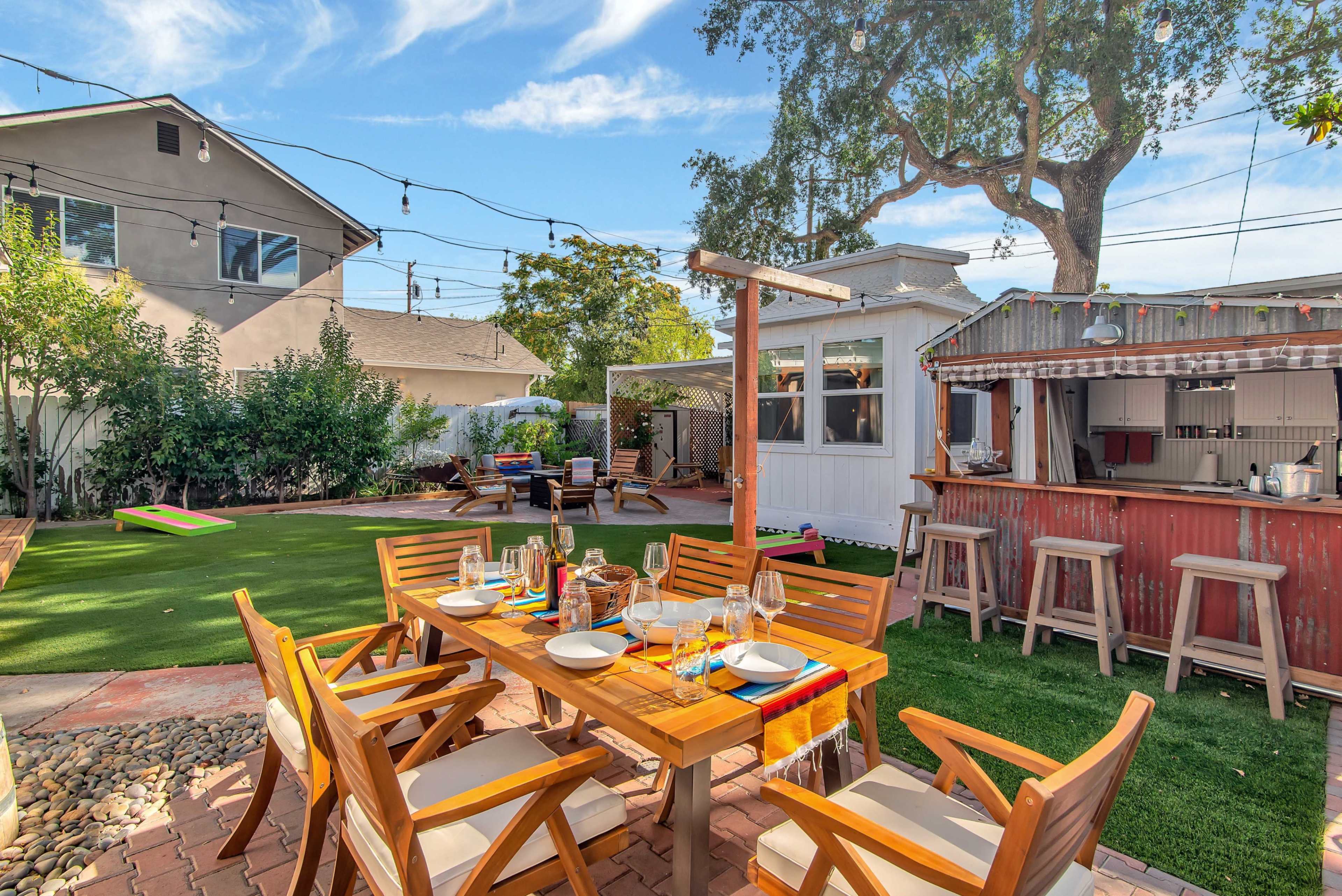 A wooden dining table is set for a meal in a backyard with a green lawn, surrounded by trees and various seating areas.