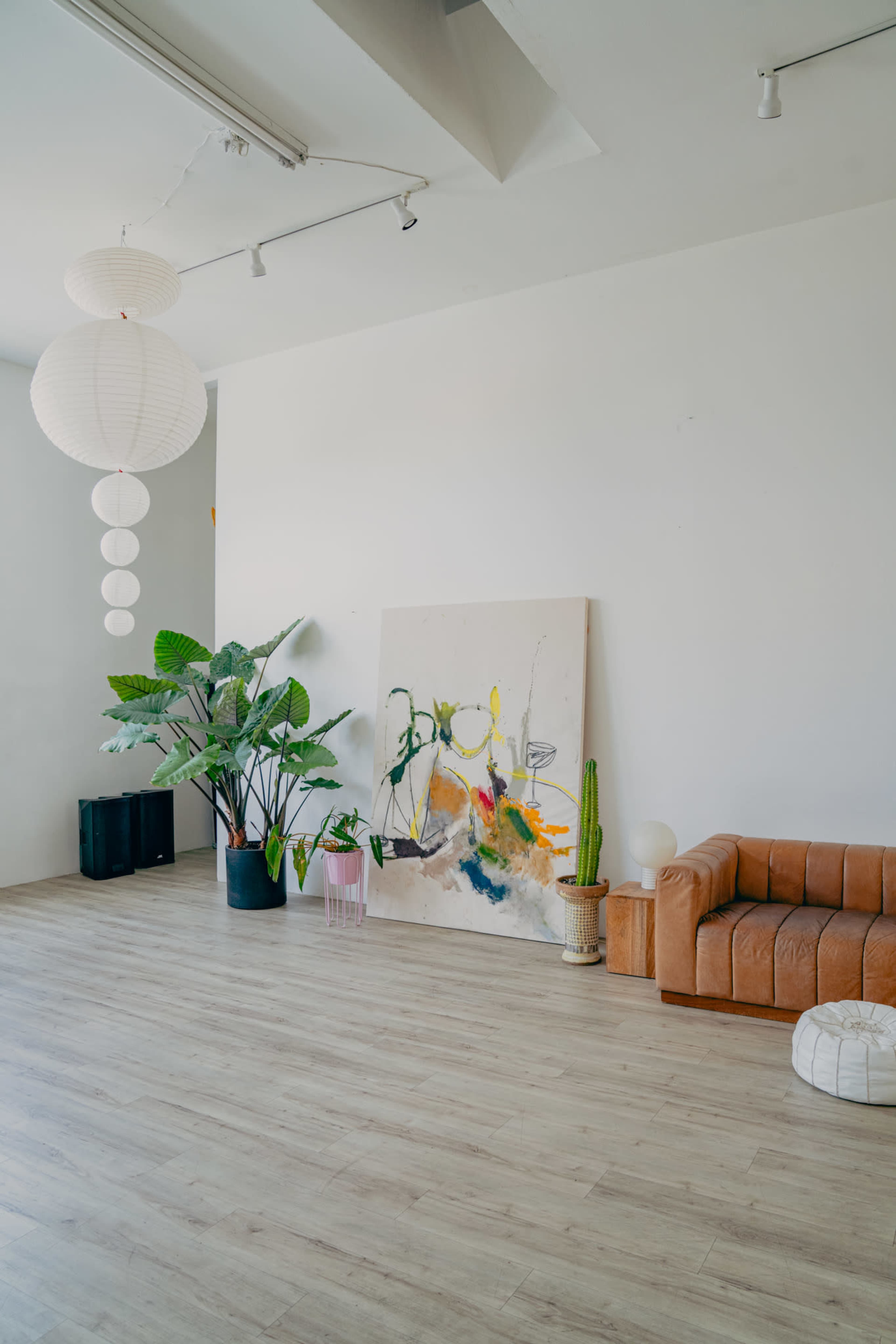 The image shows a spacious room with light wooden flooring, a large abstract painting on the wall, a brown leather couch, potted plants, and decorative lighting.
