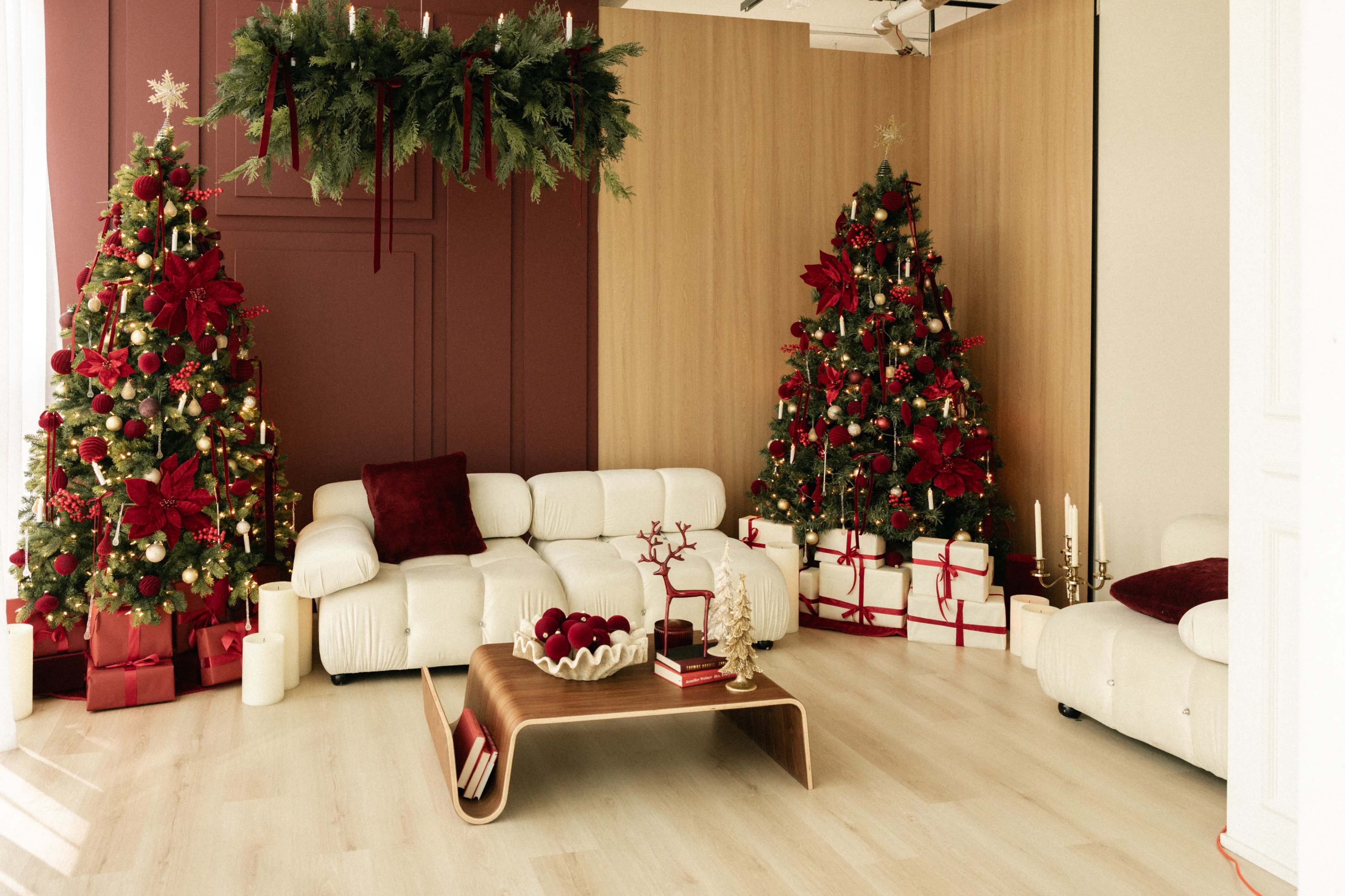 The image shows a modern living room decorated for the holidays, featuring two Christmas trees adorned with red and gold ornaments, white furniture, and a coffee table with decorative items.