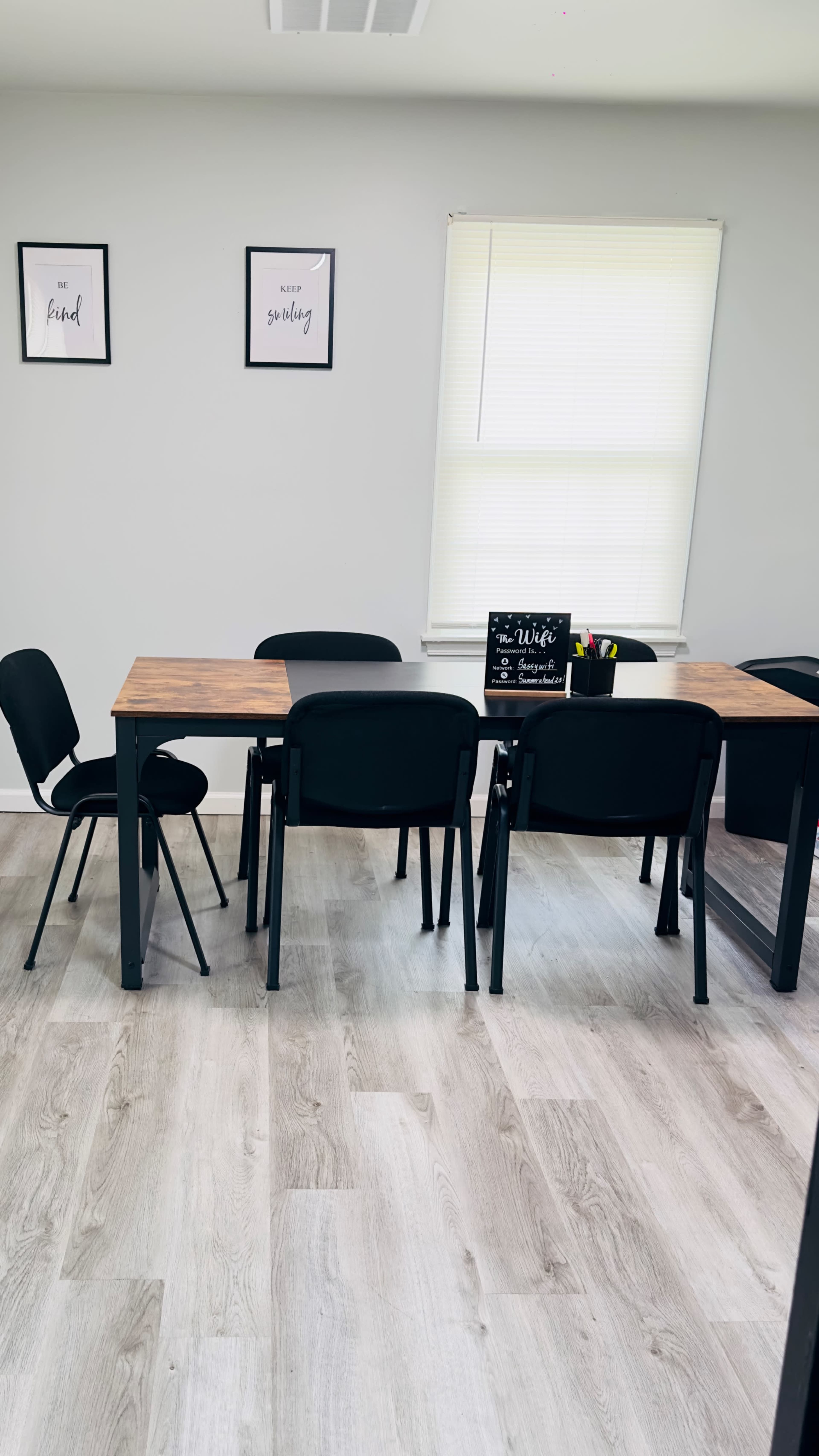 A simple dining area features a wooden table surrounded by black chairs, with framed quotes on the walls and a window letting in natural light.